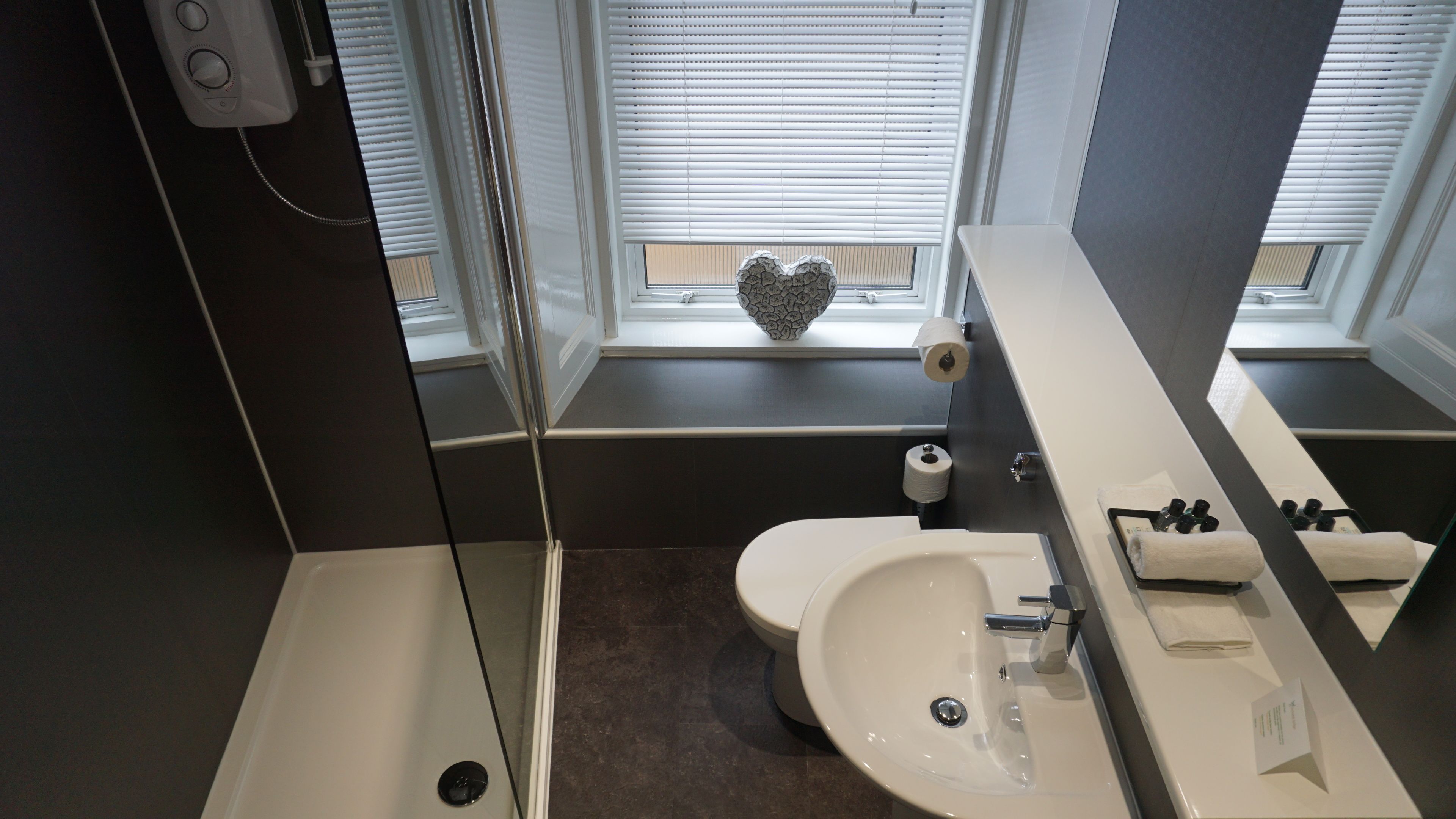 A modern bathroom with a bathtub, toilet, and sink. There are windows with blinds, a heart-shaped decoration on the windowsill, and a towel rack with towels.