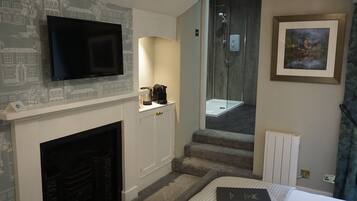 A hotel room with a flat-screen TV mounted above a fireplace, a shower area with a glass door, and a framed picture on the wall.
