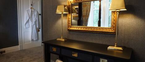 A dark-colored vanity with two lamps, a large ornate mirror, and a robe hanging on a door.