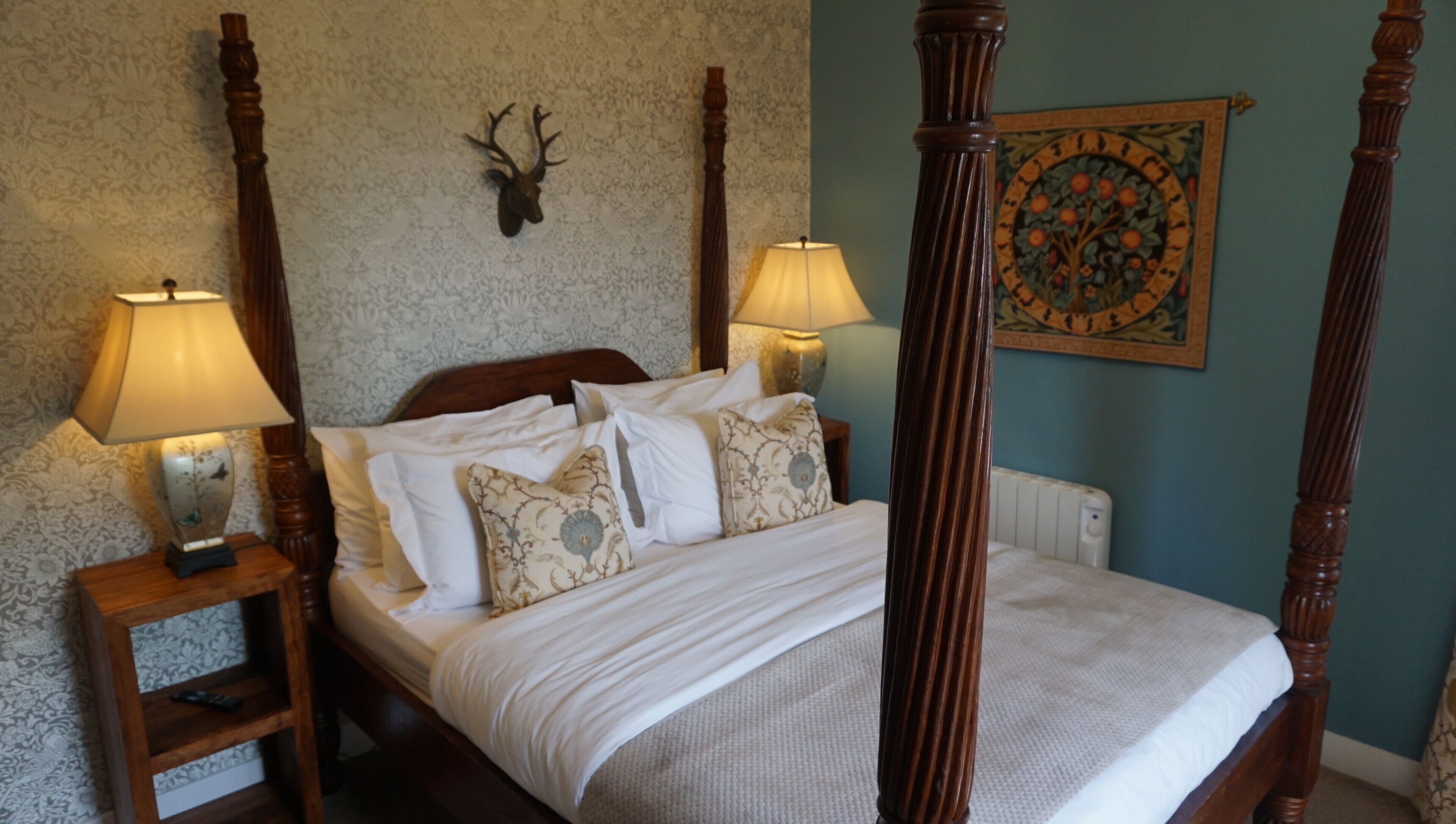 A four-poster bed with white linens, two bedside lamps, a wall-mounted deer head, and a framed floral artwork.