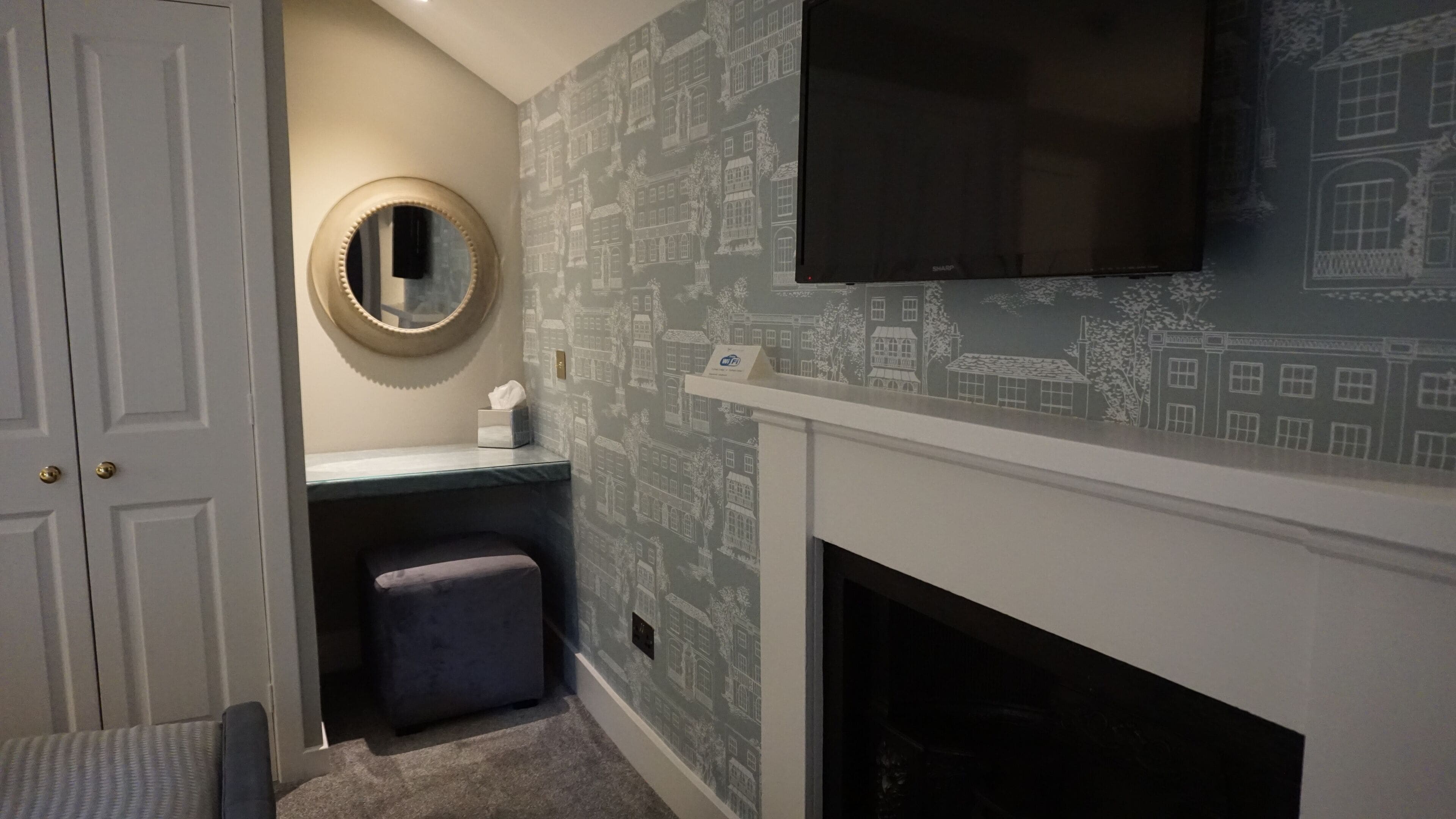 A bathroom with a vanity, a round mirror, a television mounted above the fireplace, and patterned wallpaper.