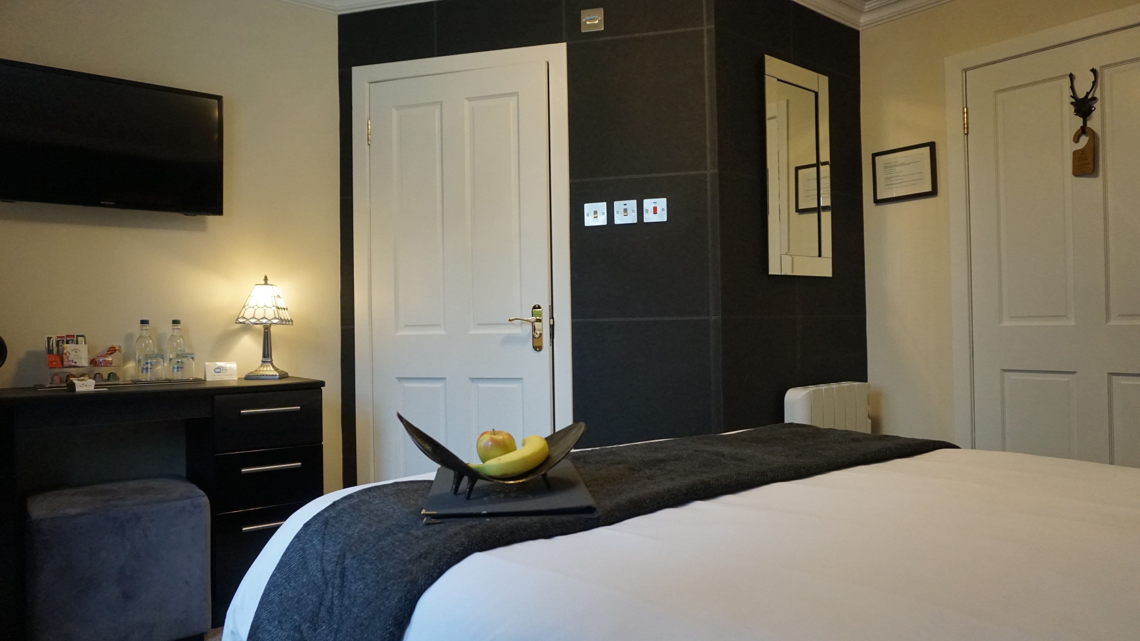 A hotel room with a bed, a desk with a lamp, a mirror, and a fruit bowl on the bed.