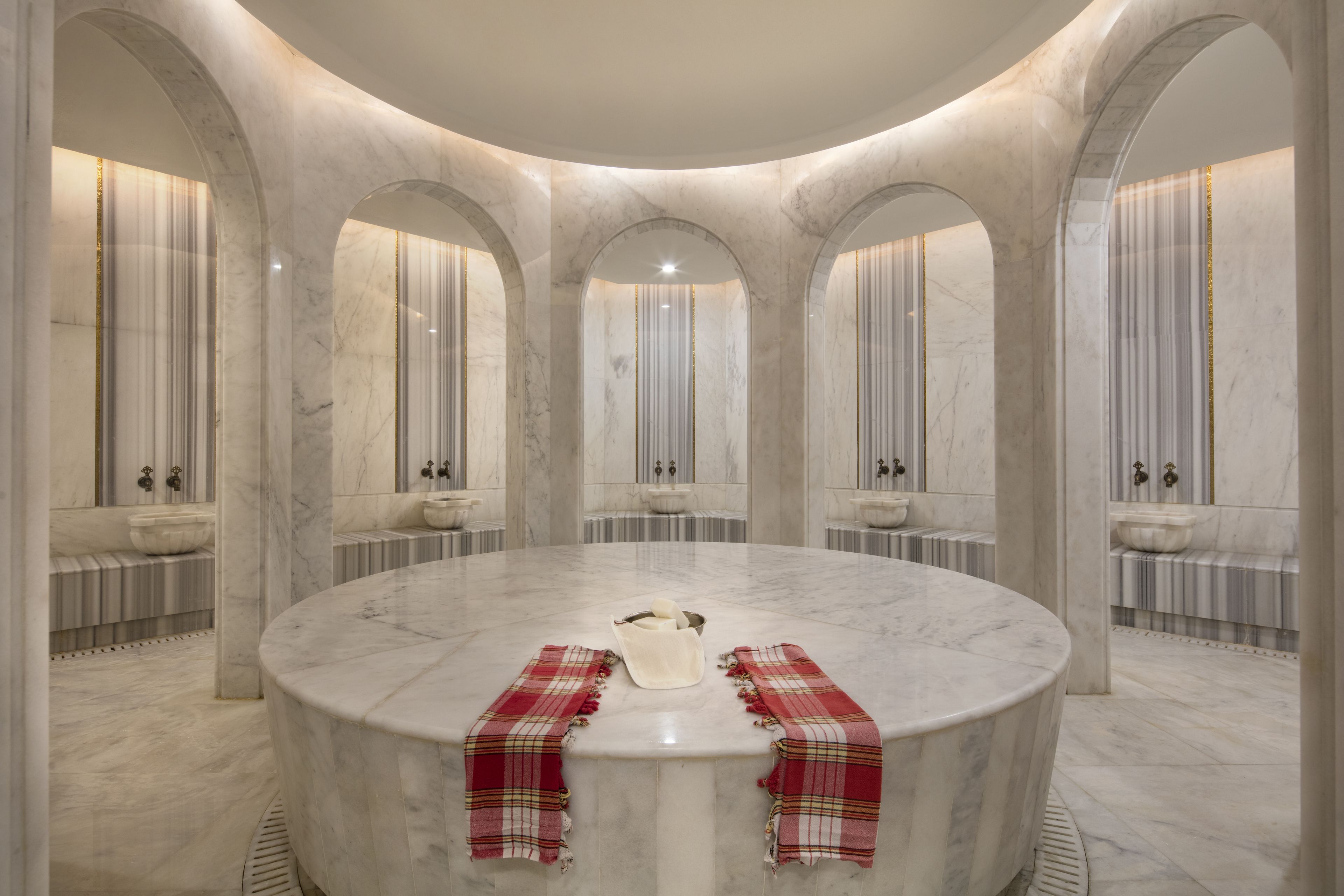 A circular marble-topped table with a bowl and cloths in the center, surrounded by arched marble walls and sinks.