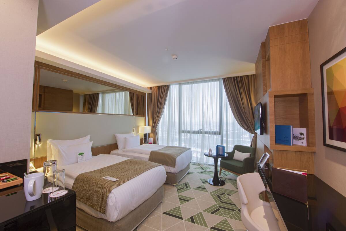 Une chambre d’hôtel avec deux lits, un bureau avec un ordinateur, une chaise, une petite table avec une lampe et une grande fenêtre avec des rideaux.