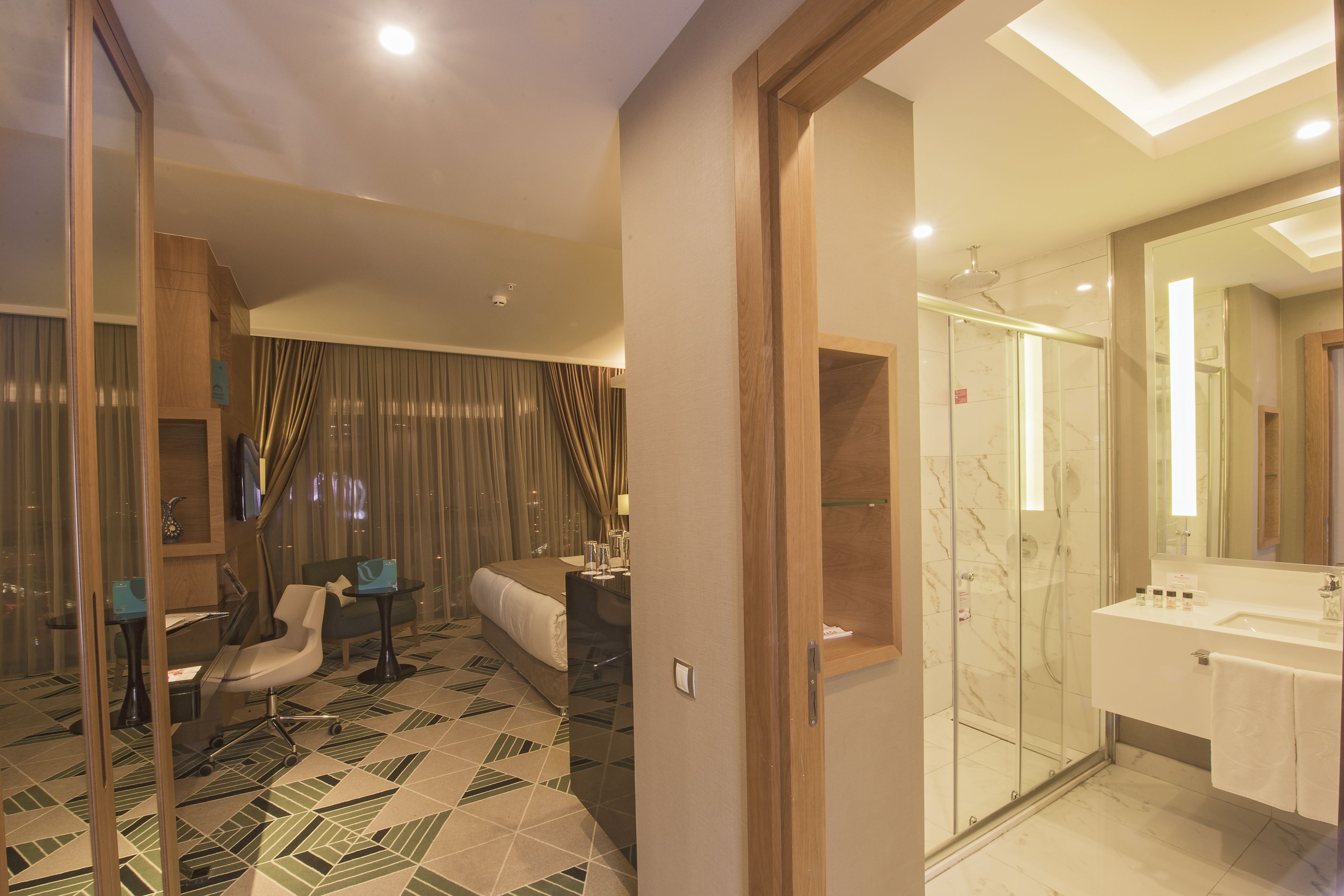 A modern hotel room with a bed, a desk, a chair, a glass-enclosed shower, and a bathroom with a sink and mirror.