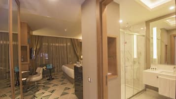 A modern hotel room with a bed, a desk, a chair, a glass-enclosed shower, and a bathroom with a sink and mirror.