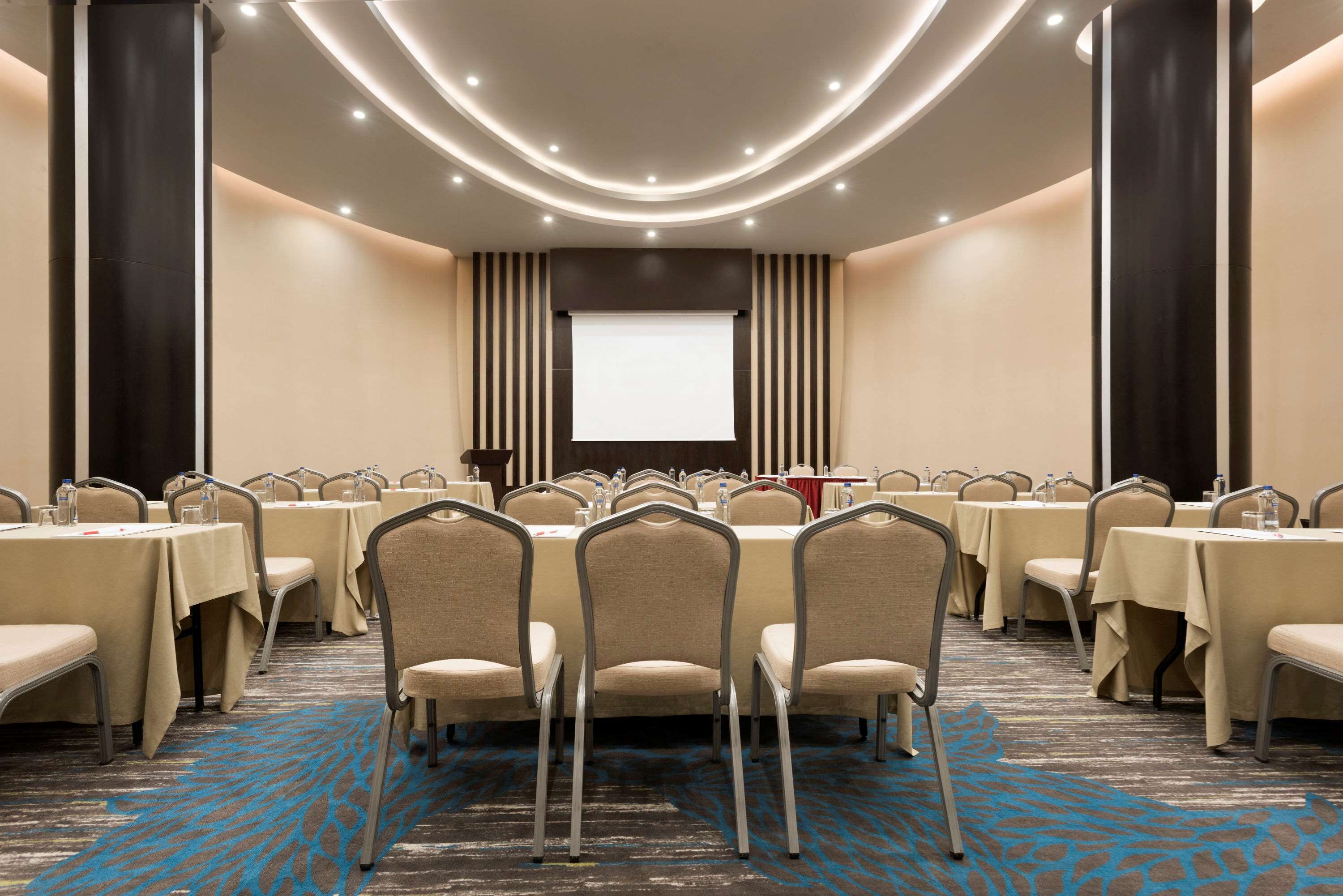 Une salle de conférence dotée de plusieurs tables et chaises, d’un écran de projection et d’un sol recouvert de moquette.
