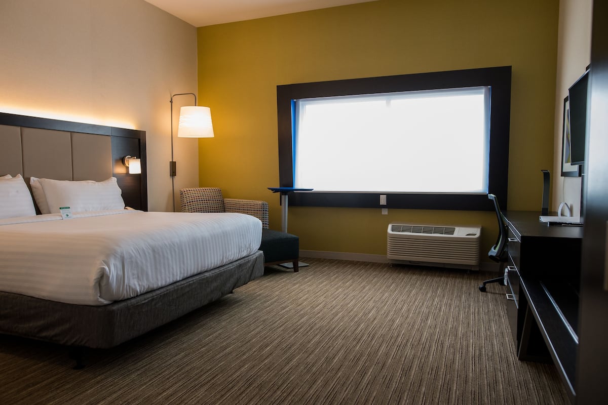 A hotel room with a bed, bedside lamp, window, air conditioning unit, and a desk with a computer.