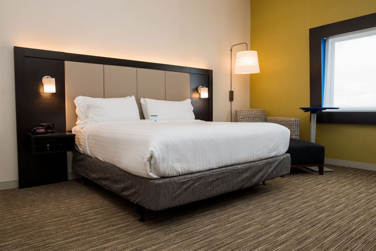 A neatly made bed with a headboard, a bedside table with a lamp, and a window with blinds.