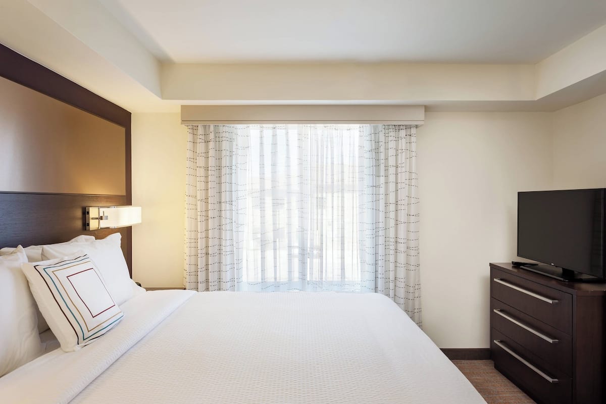 A hotel room with a bed, a TV on a dresser, and a window with sheer curtains.