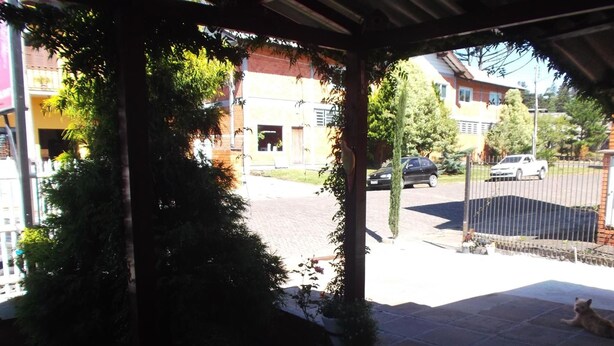 A covered patio area with a tiled floor, surrounded by greenery and a view of a residential street with parked cars and houses. - Pousada Roses Canela (Canela)