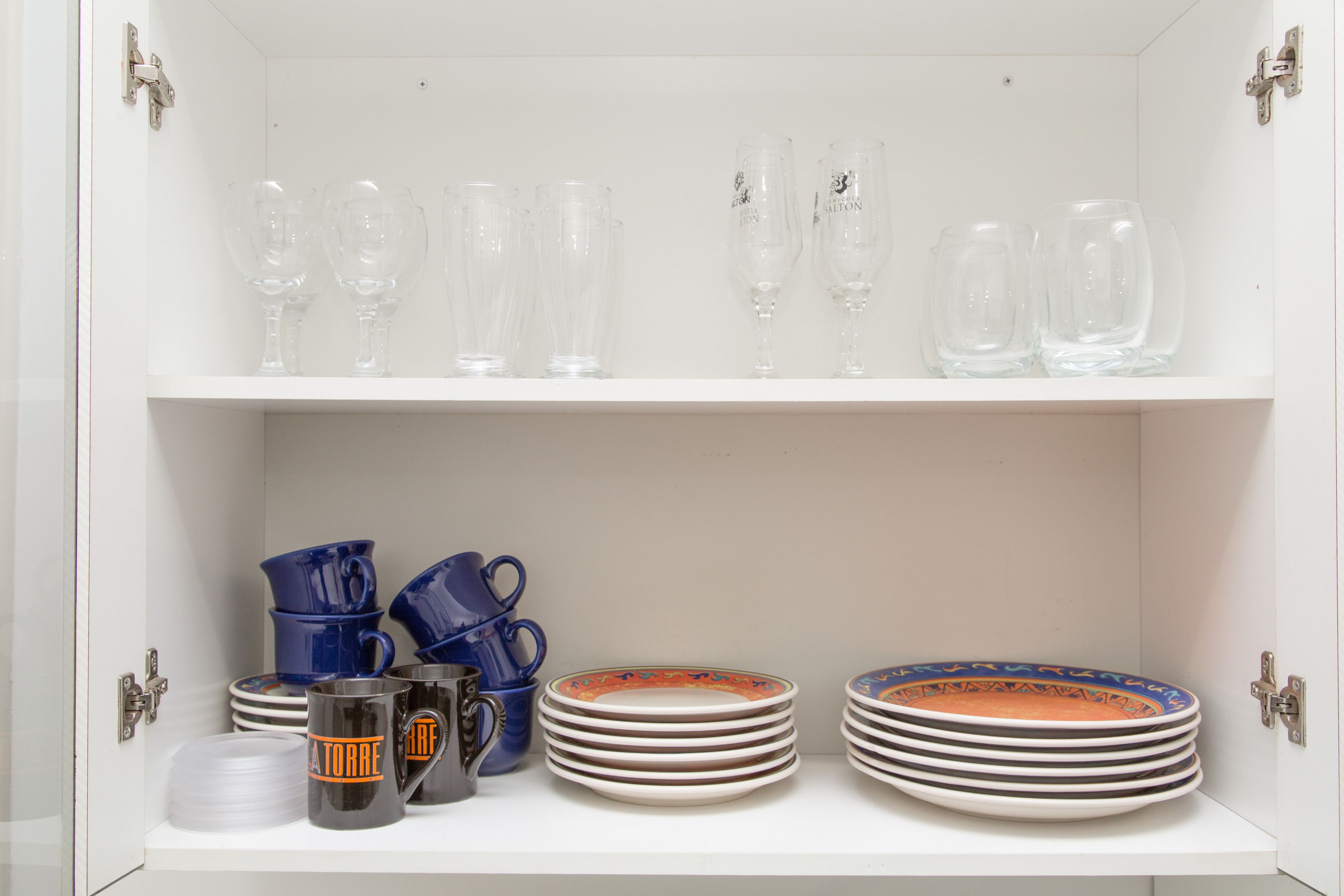 A white cabinet with shelves containing clear wine glasses, blue pitchers, and stacked plates with colorful rims.