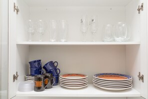 A white cabinet with shelves containing clear wine glasses, blue pitchers, and stacked plates with colorful rims. - Apartamento Redenção (Porto Alegre)