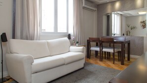 Una sala de estar moderna con un sofá blanco, una mesa de comedor con sillas, una mesita auxiliar con una planta en maceta y un amplio ventanal con cortinas.