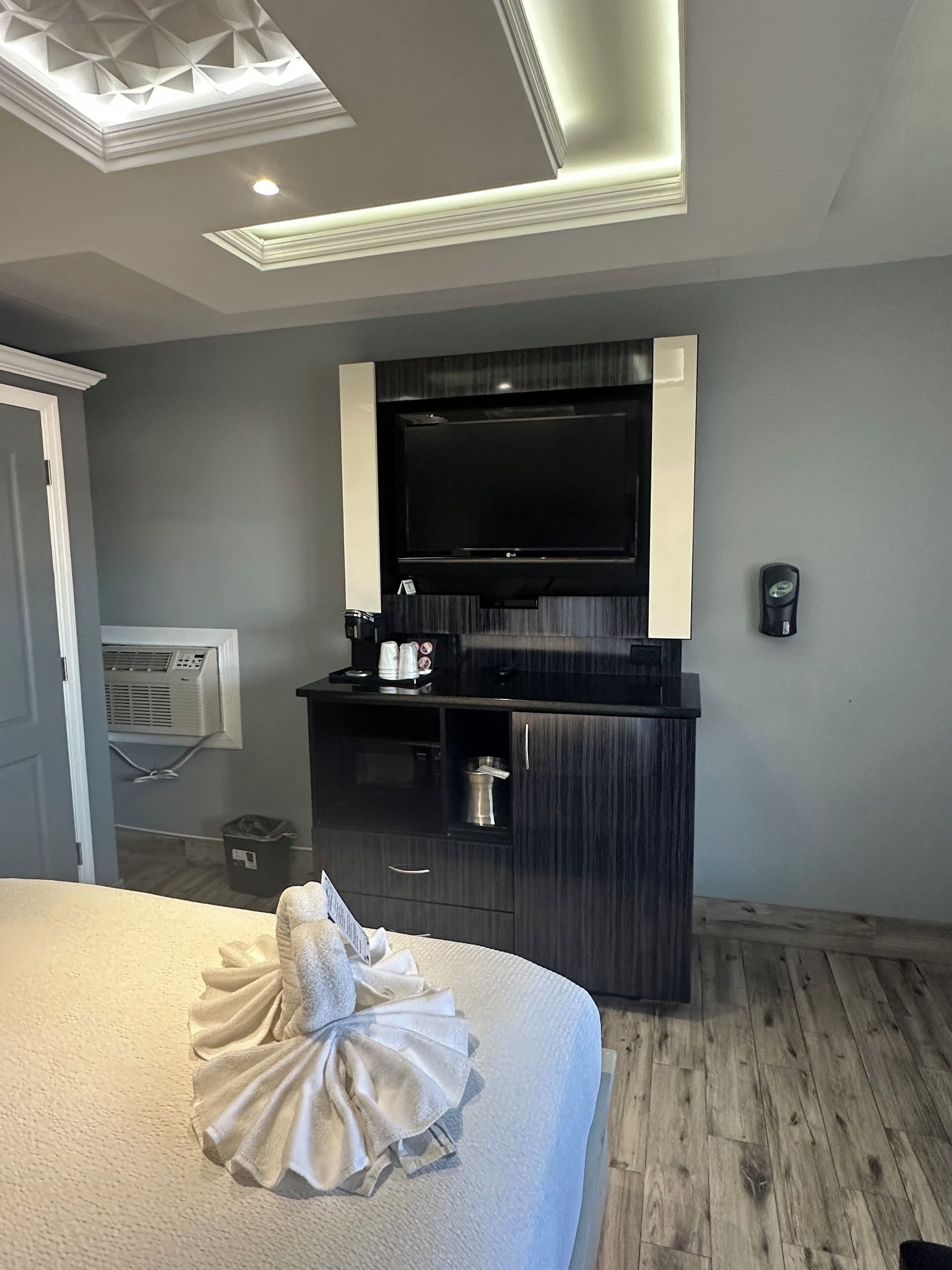 A hotel room with a large bed, a flat-screen TV mounted on the wall, a minibar, and a ceiling light fixture.
