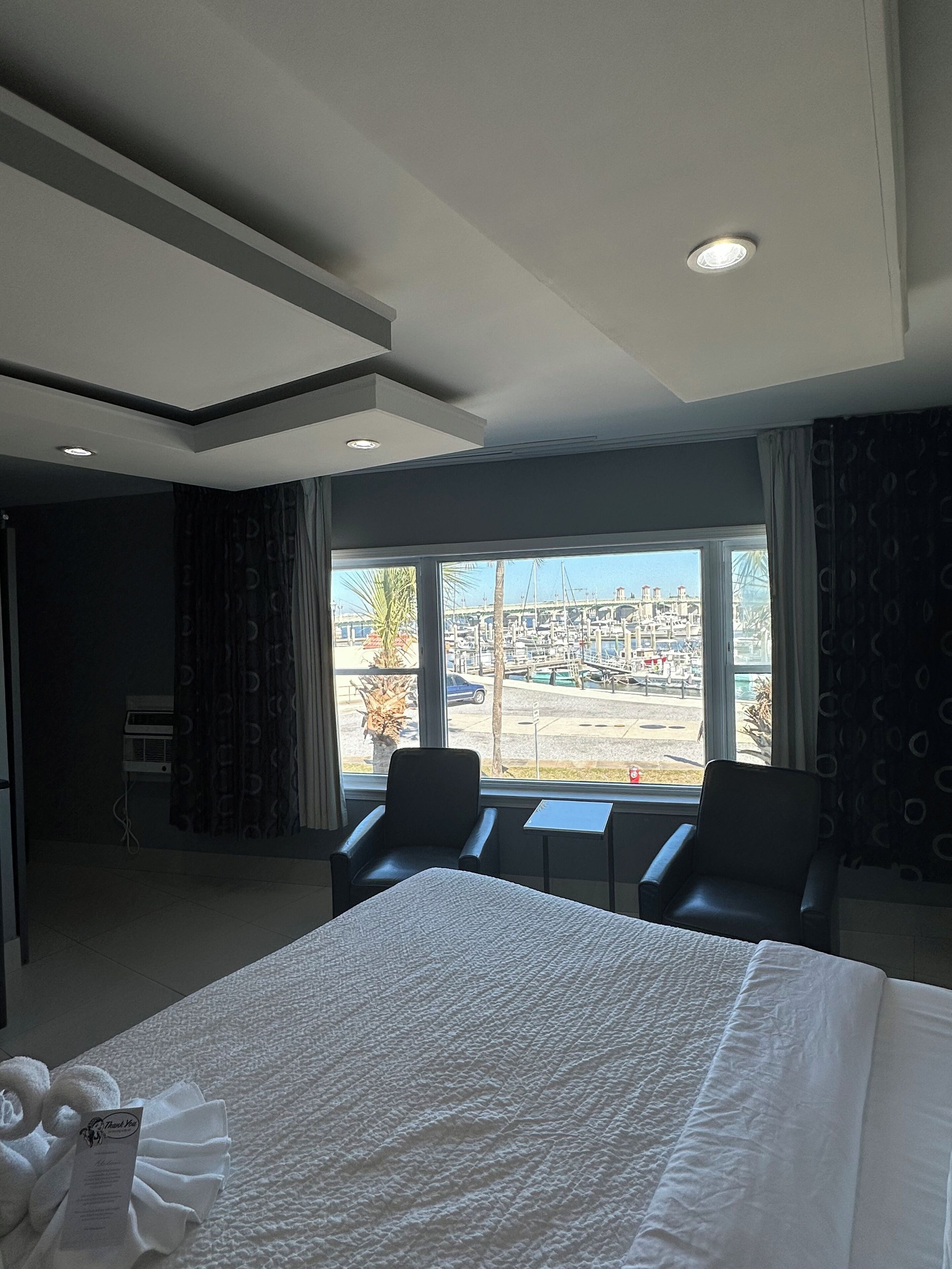 Habitación de hotel con una cama grande, dos sillas, una mesita y vistas a un puerto deportivo y al paisaje urbano a través de la ventana.