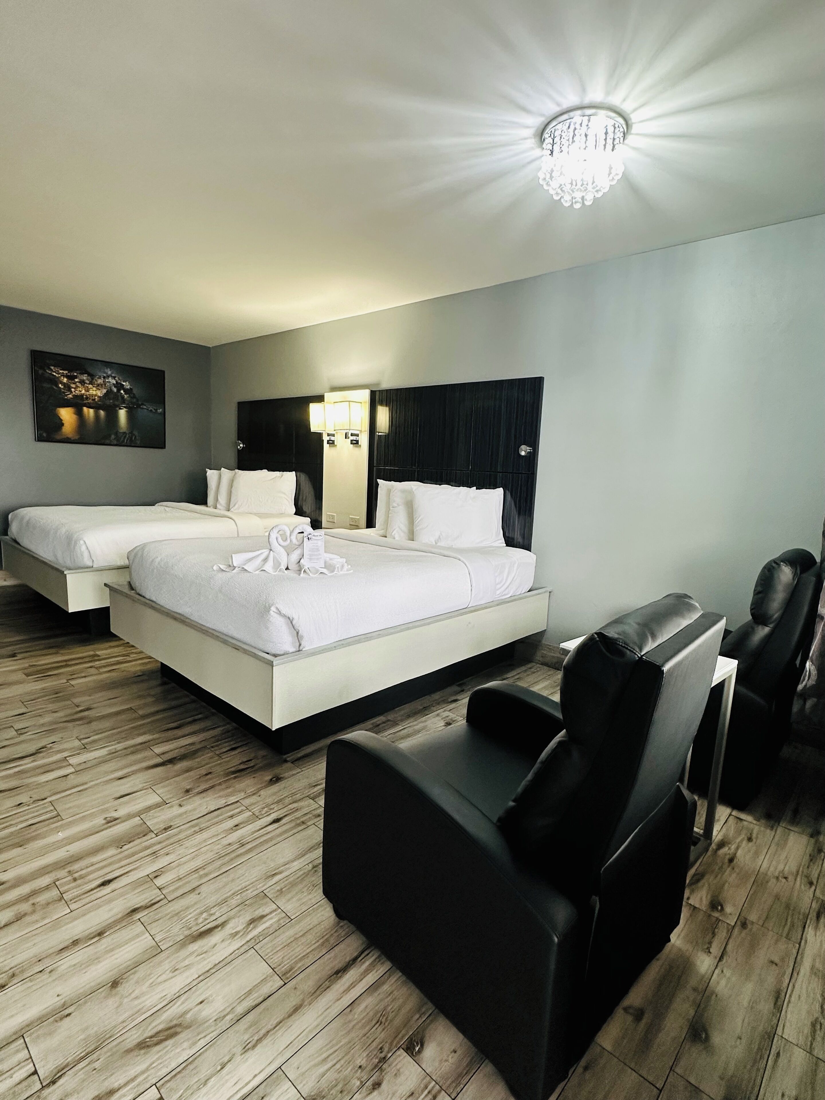 A hotel room with two beds, a black leather chair, a wooden floor, a chandelier, and a wall-mounted TV.