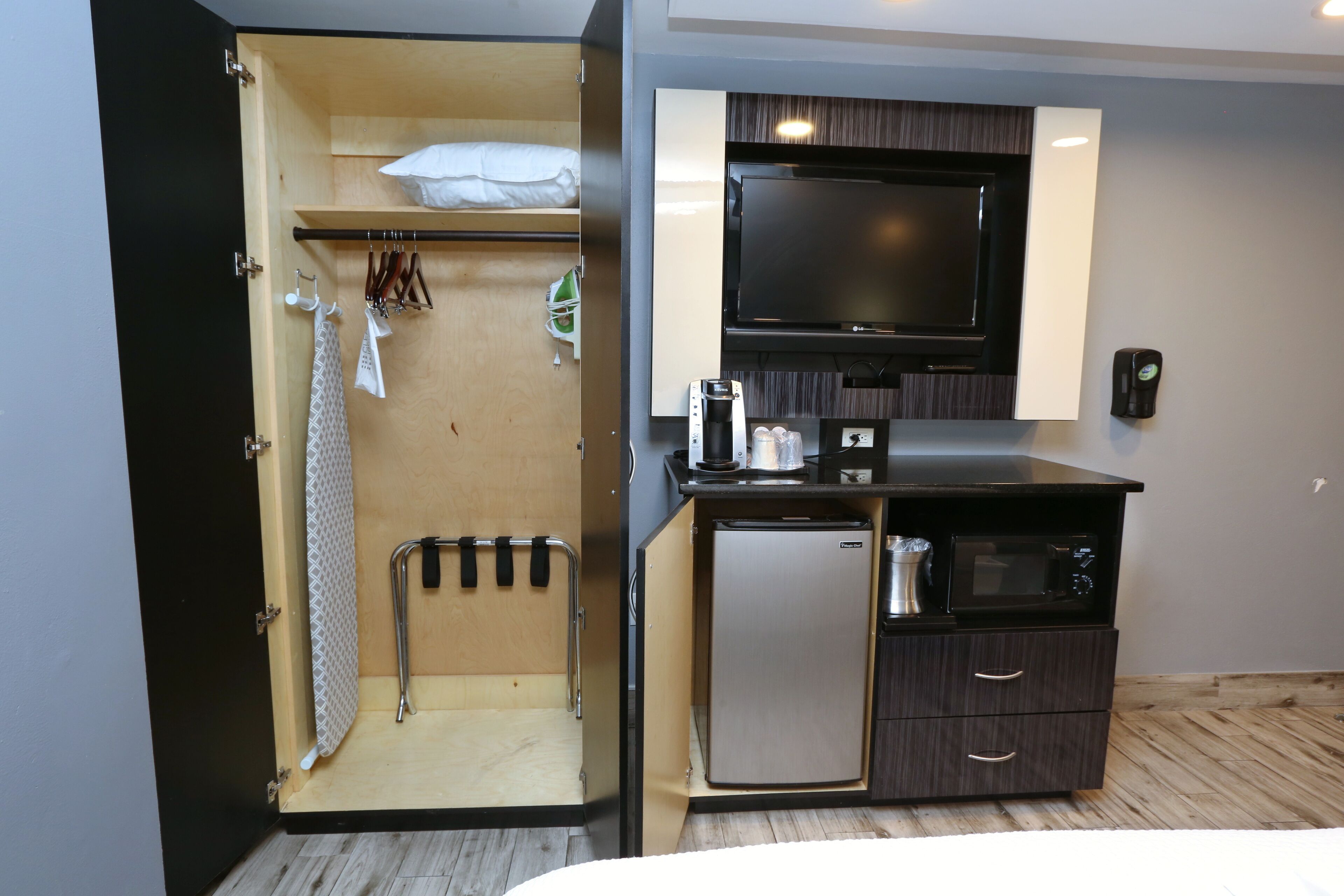 A compact hotel room with a kitchenette, a TV, a small refrigerator, and a wardrobe with hanging clothes.