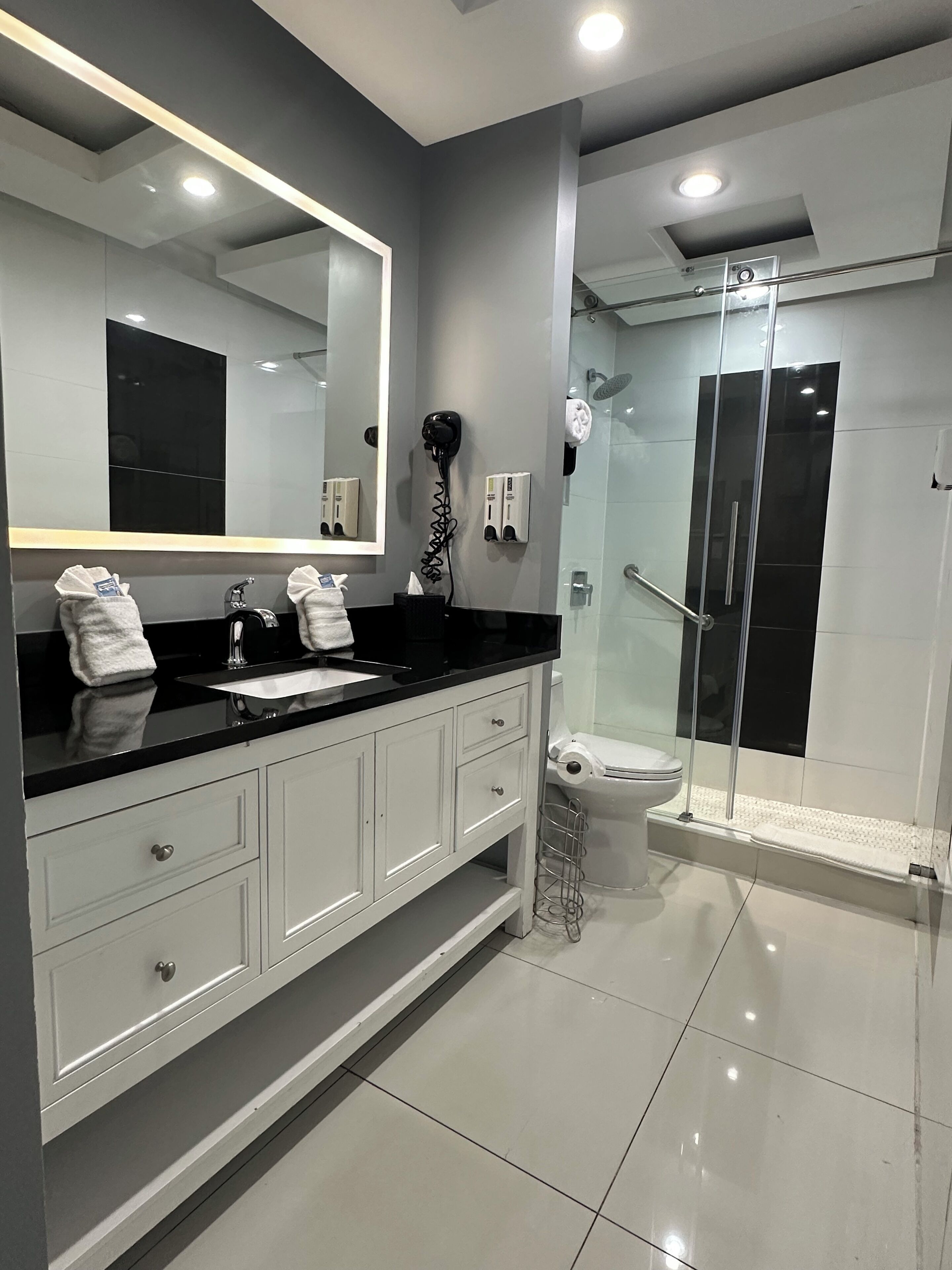 Un baño moderno con encimera negra, muebles blancos, un espejo grande y una cabina de ducha de cristal.