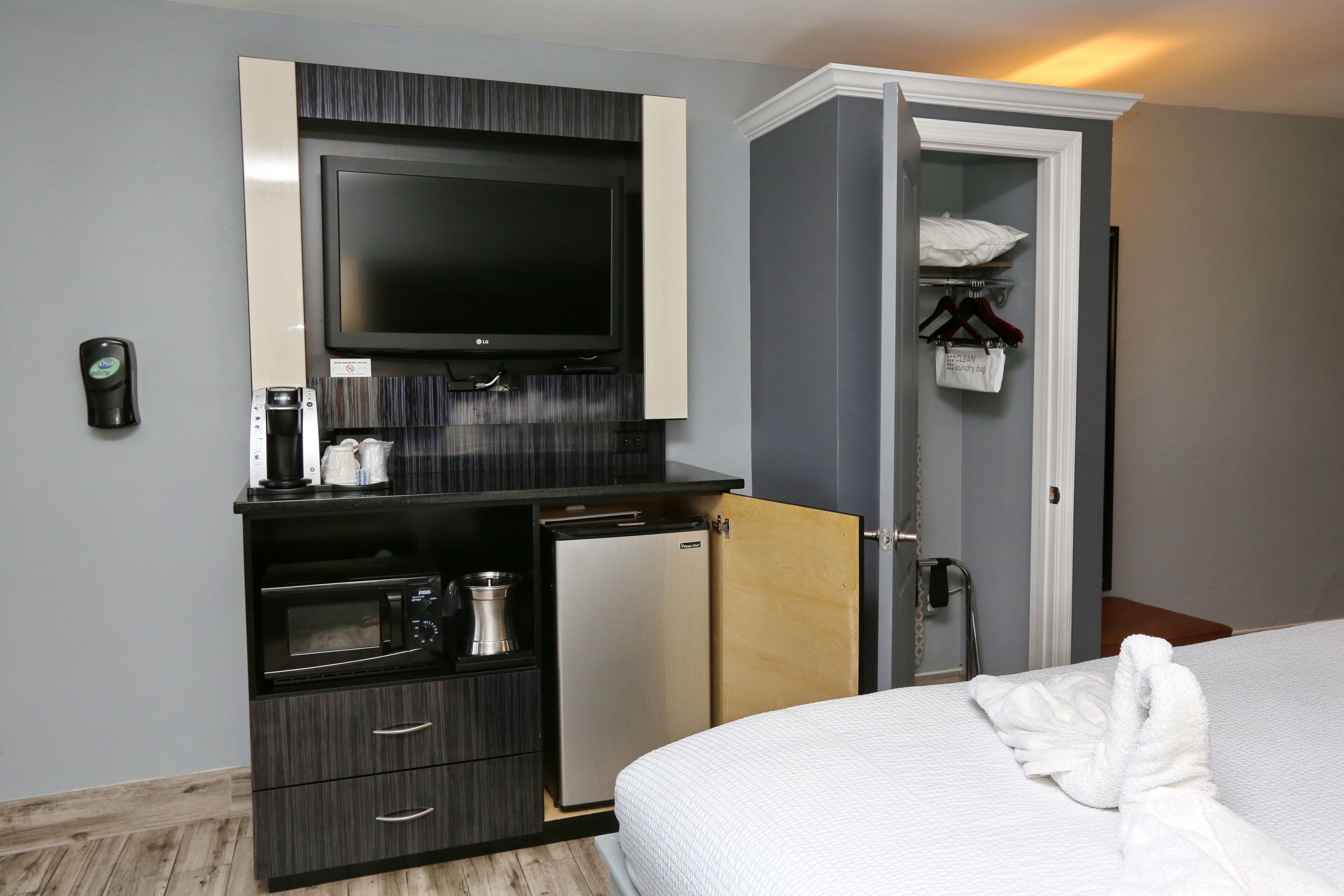 Une chambre d’hôtel comprenant un lit, un téléviseur fixé sur un meuble de couleur sombre, un petit réfrigérateur, un four à micro-ondes et une porte en bois donnant sur un placard où se trouve un sac suspendu.