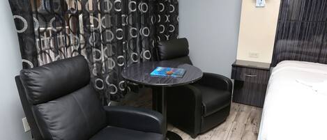 A modern room with a black leather sofa, a small round table with a magazine, a bed with a white duvet, and a wall-mounted lamp.