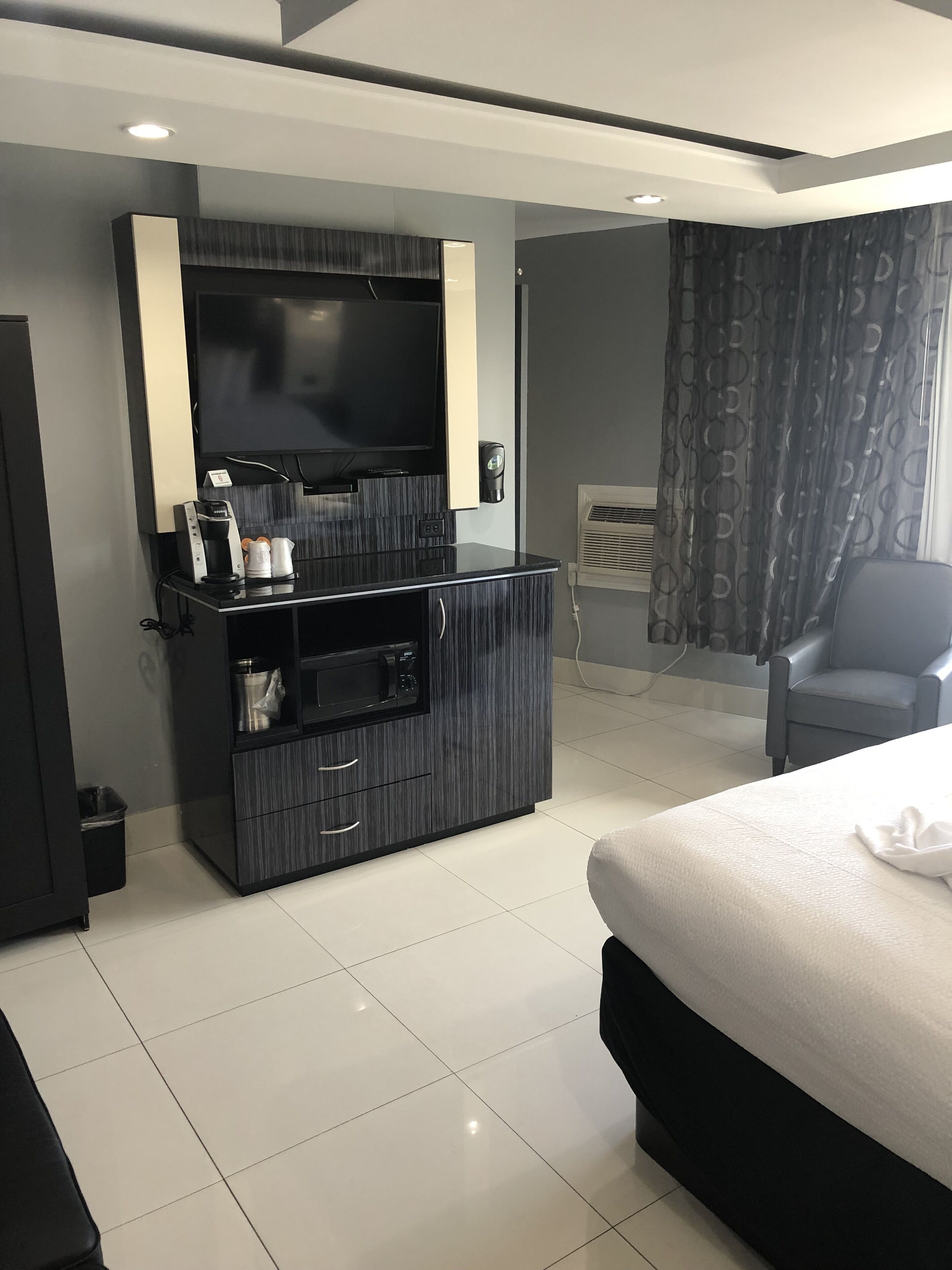 Una habitación de hotel moderna con una cama grande, un televisor de pantalla plana montado en un mueble de madera oscura, una cafetera y un pequeño cubo de basura.