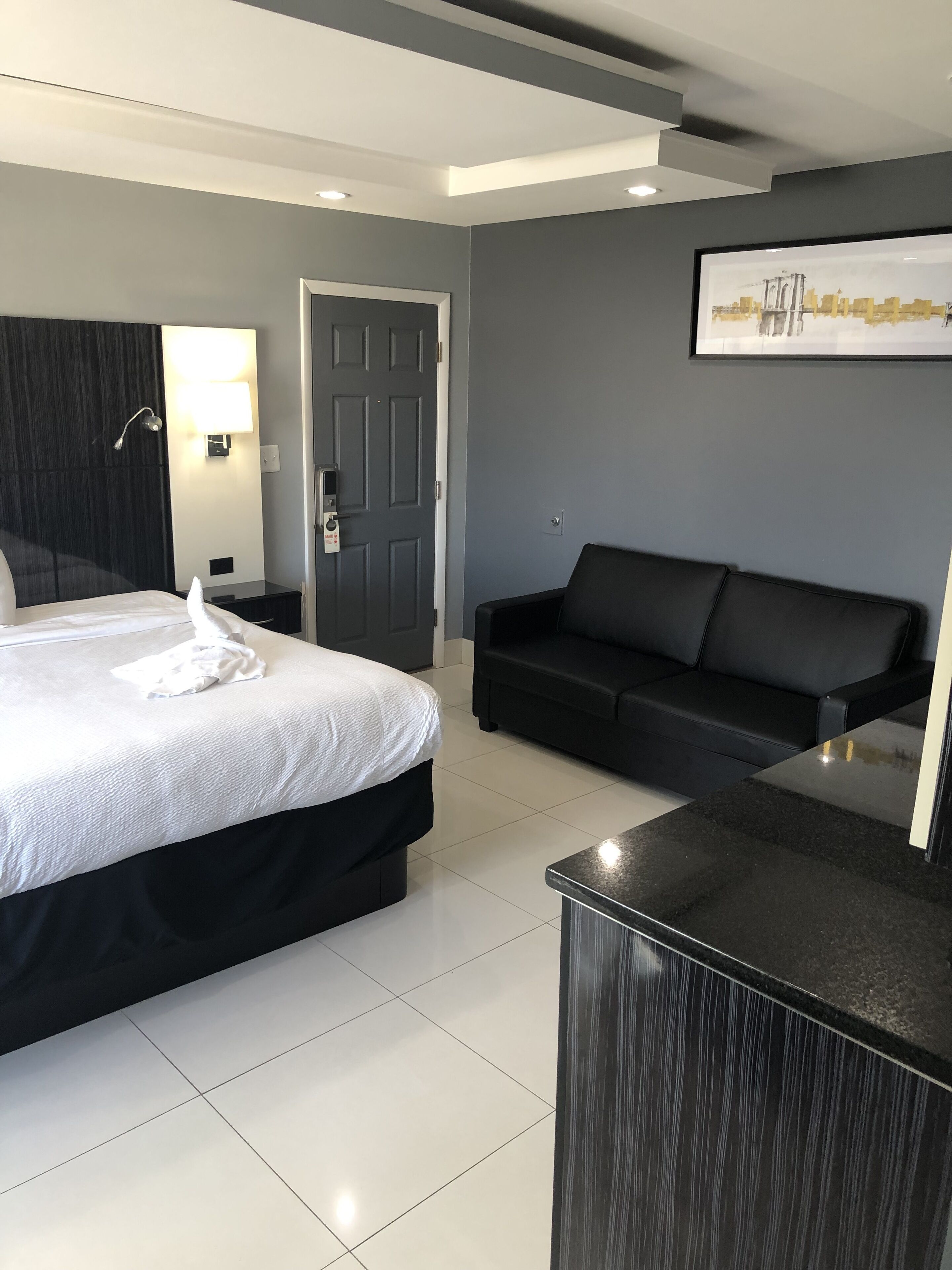 A modern hotel room with a bed, a black sofa, a desk, and a framed picture on the wall.