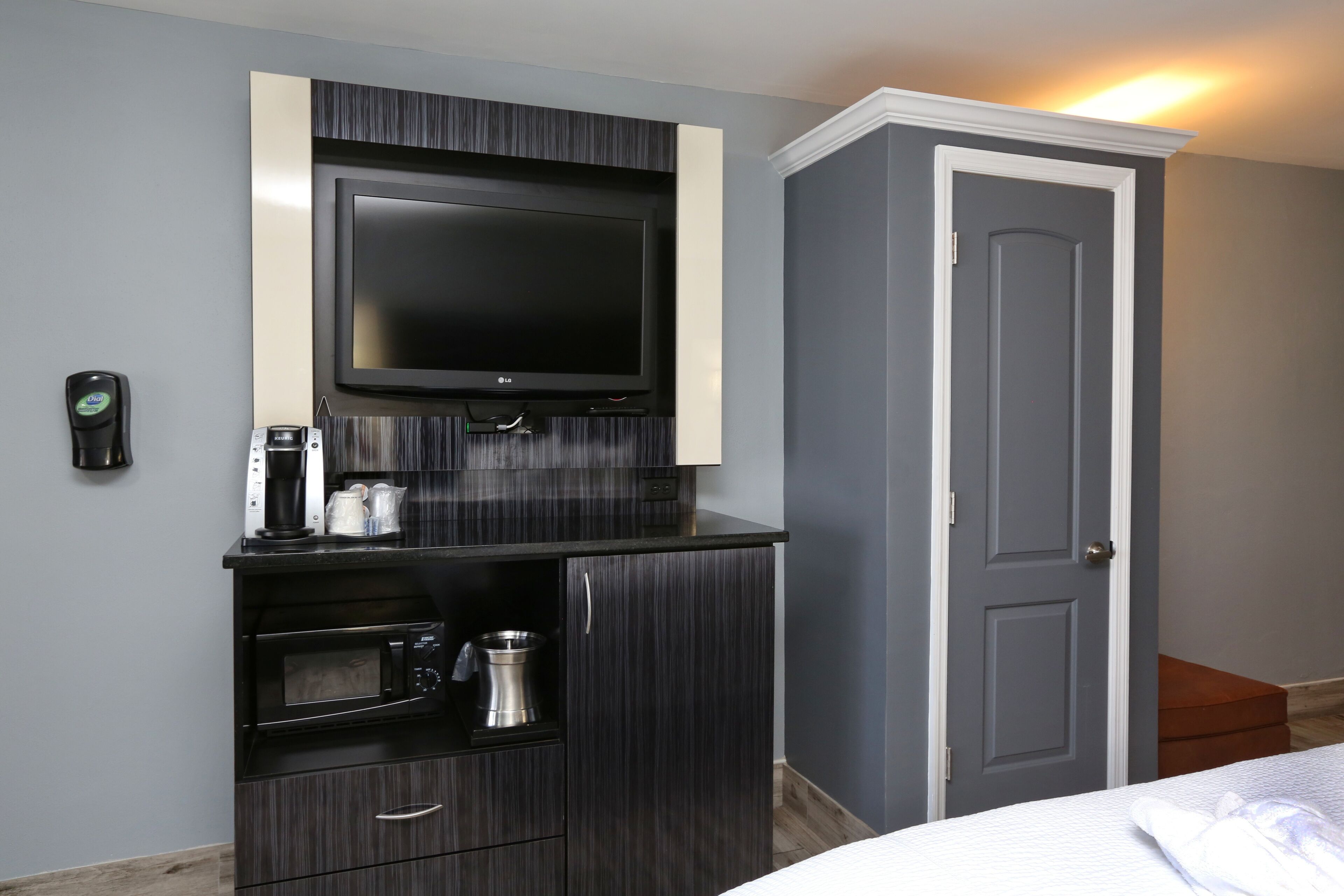 A compact hotel room with a flat-screen TV mounted on a dark wood cabinet, a coffee maker, a microwave, and a small storage cabinet.