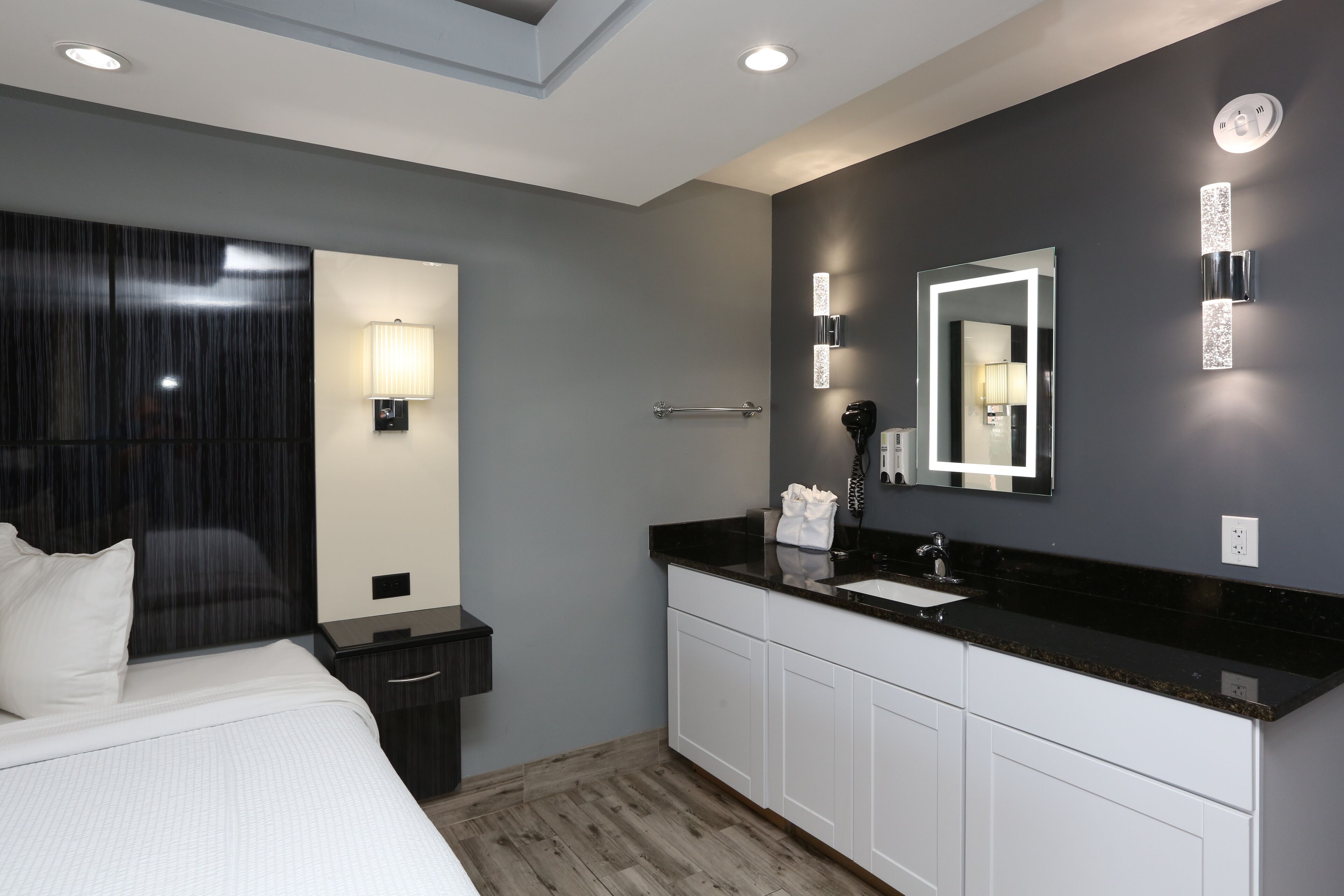 A modern hotel room with a large bed, a dark headboard, a bedside table, a nightstand, a black countertop with a sink, a mirror, and a wall-mounted light fixture.