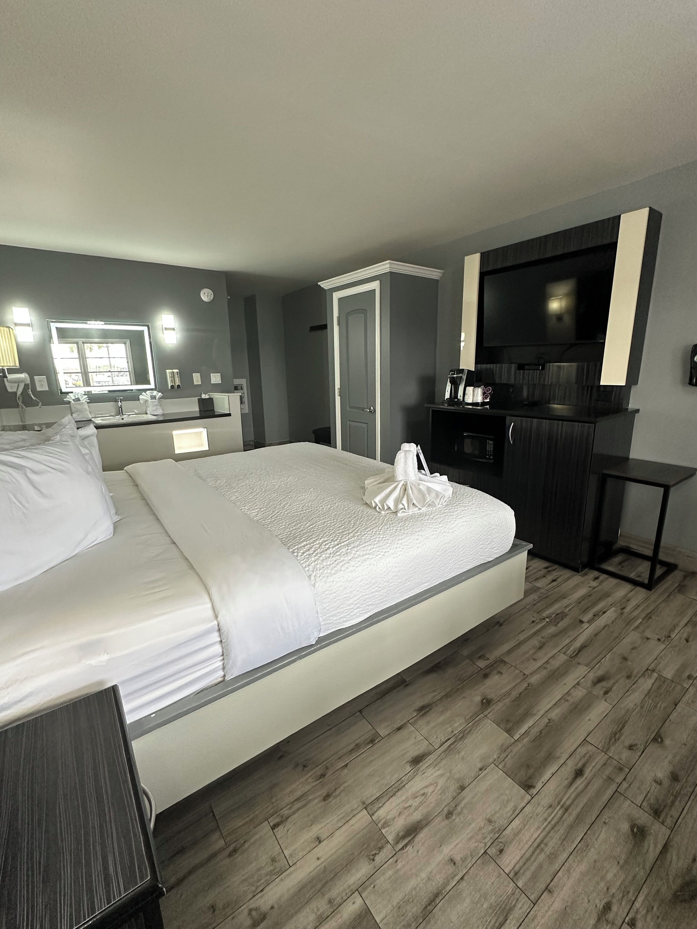 A hotel room with a large bed, a flat-screen TV, a small kitchenette, and a bathroom with a mirror and lights.