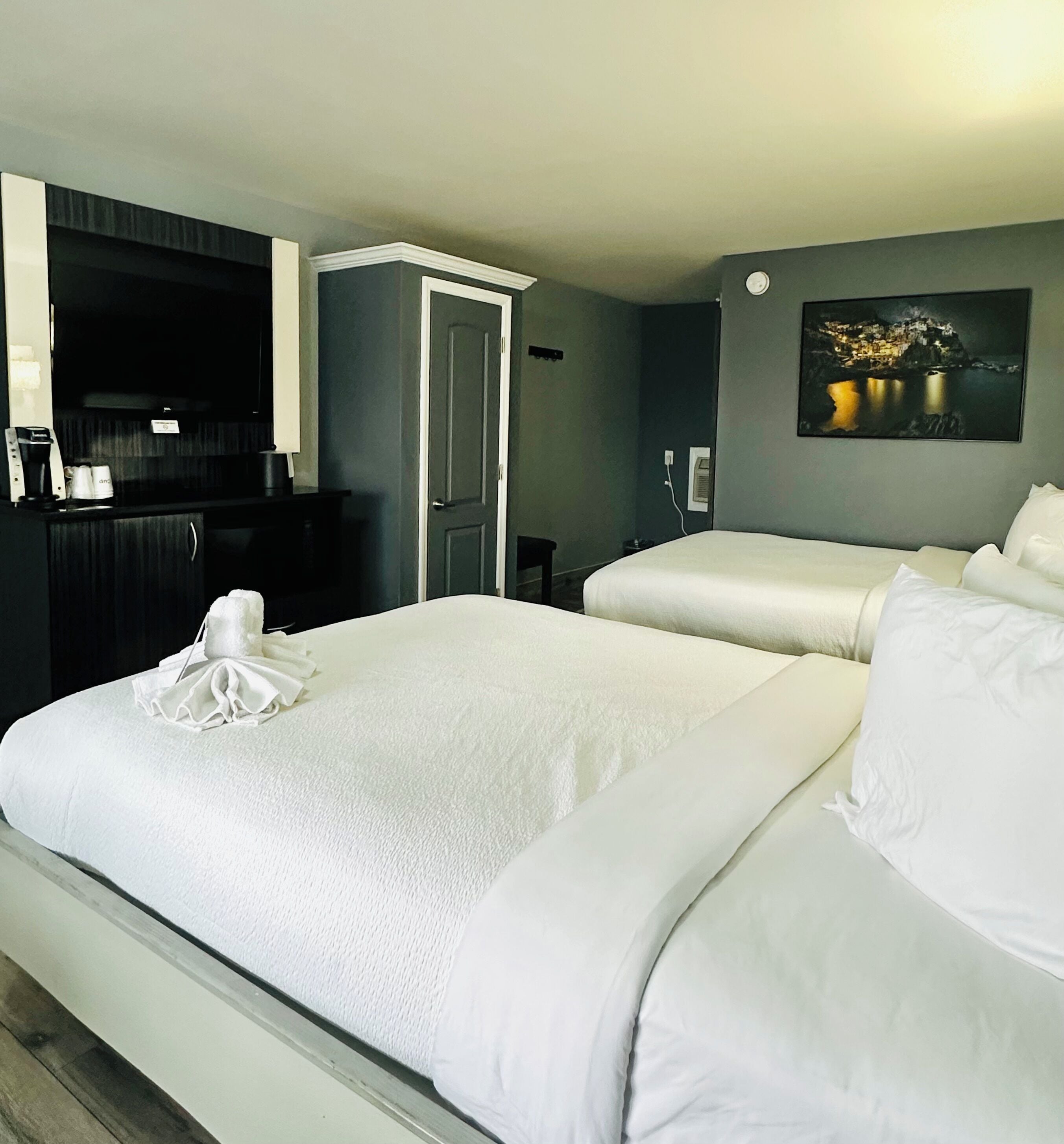 A hotel room with two beds, a dark-colored wardrobe, a small desk with a coffee maker, and a painting on the wall.