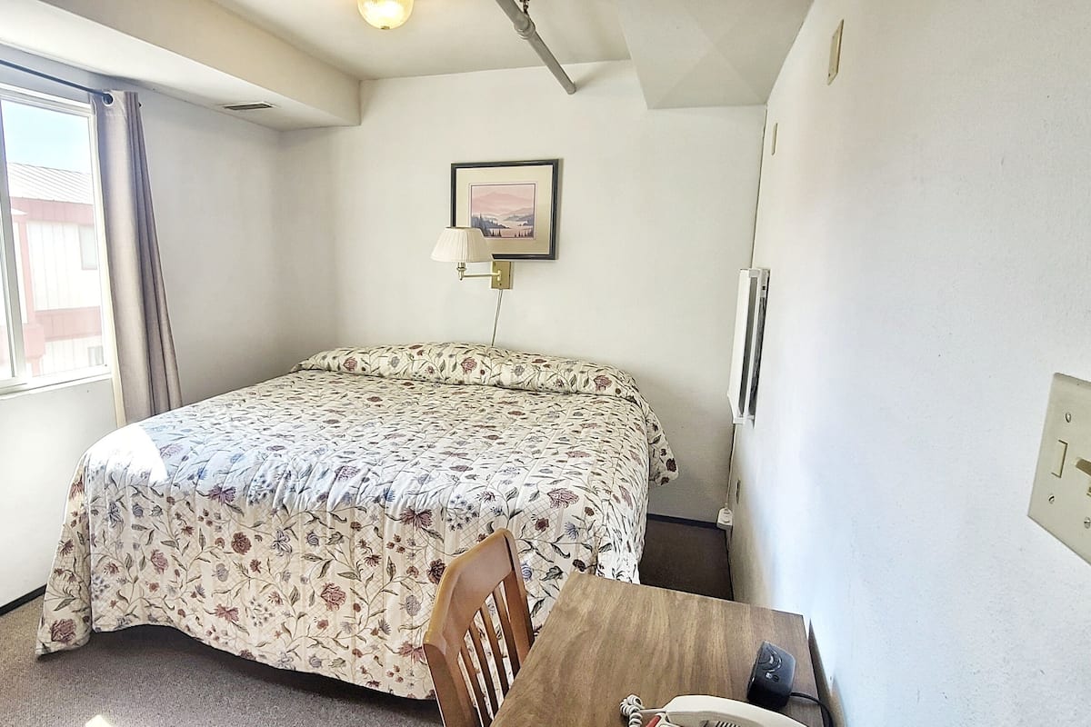 A small, neatly arranged bedroom with a bed, a bedside table, a lamp, and a window with curtains.