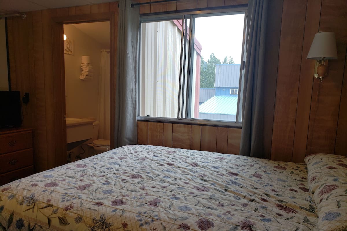 A bedroom with a floral bedspread, wooden walls, a window with curtains, a bedside lamp, and a view of a building outside.