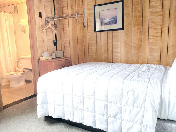 A bedroom with a wooden wall, a bed with a white quilt, a wooden dresser, a framed picture, and a bathroom with a toilet and shower curtain.