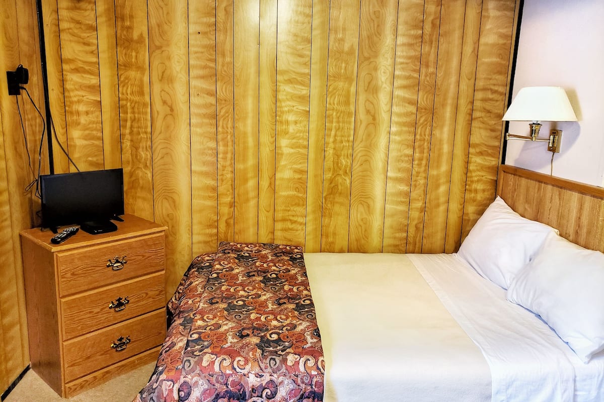 A hotel room with a wooden panel wall, a wooden dresser with drawers, a television on top, a patterned bedspread, white pillows, and a wall-mounted lamp.