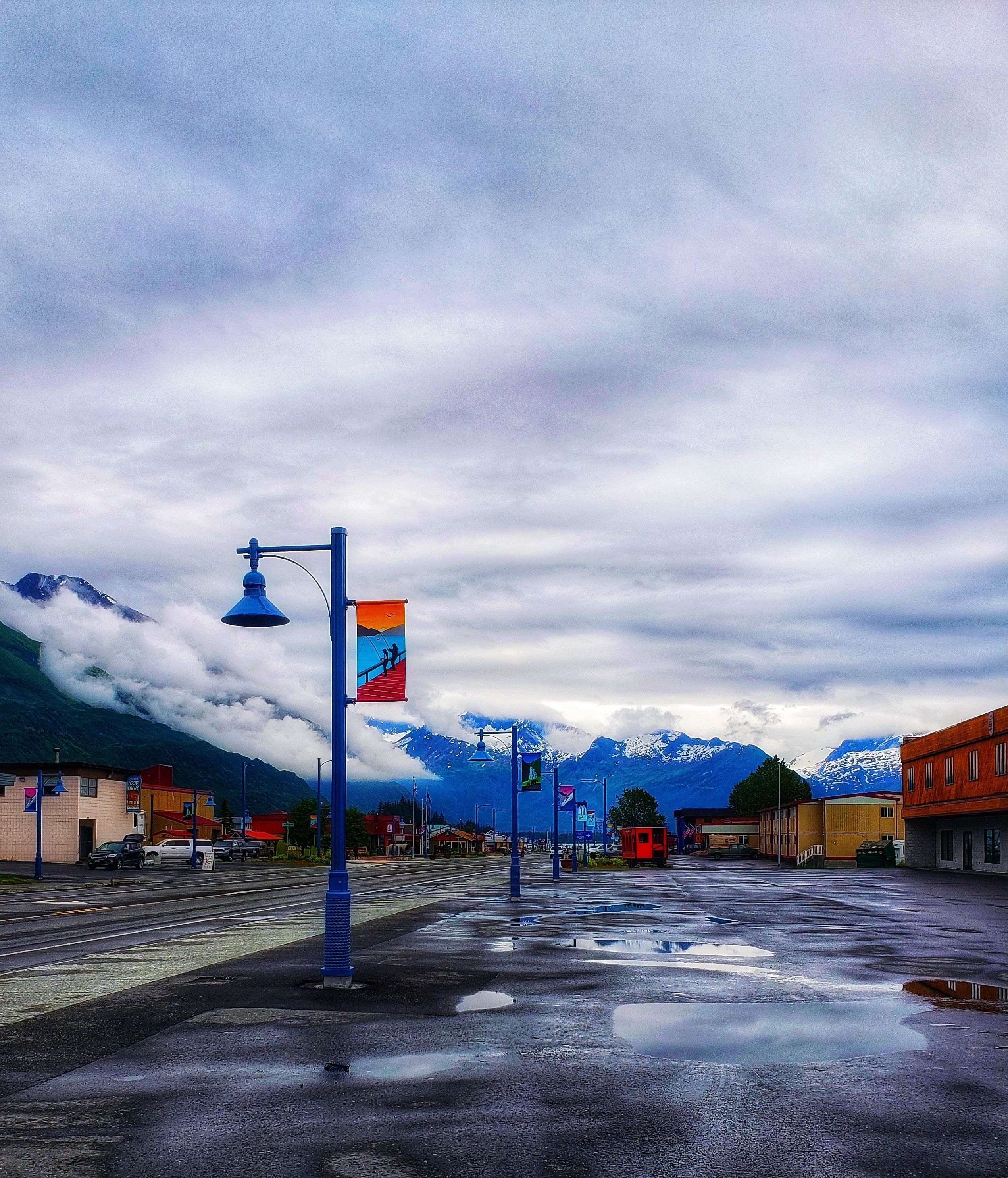 A street with buildings, a mountain range, and a cloudy sky.