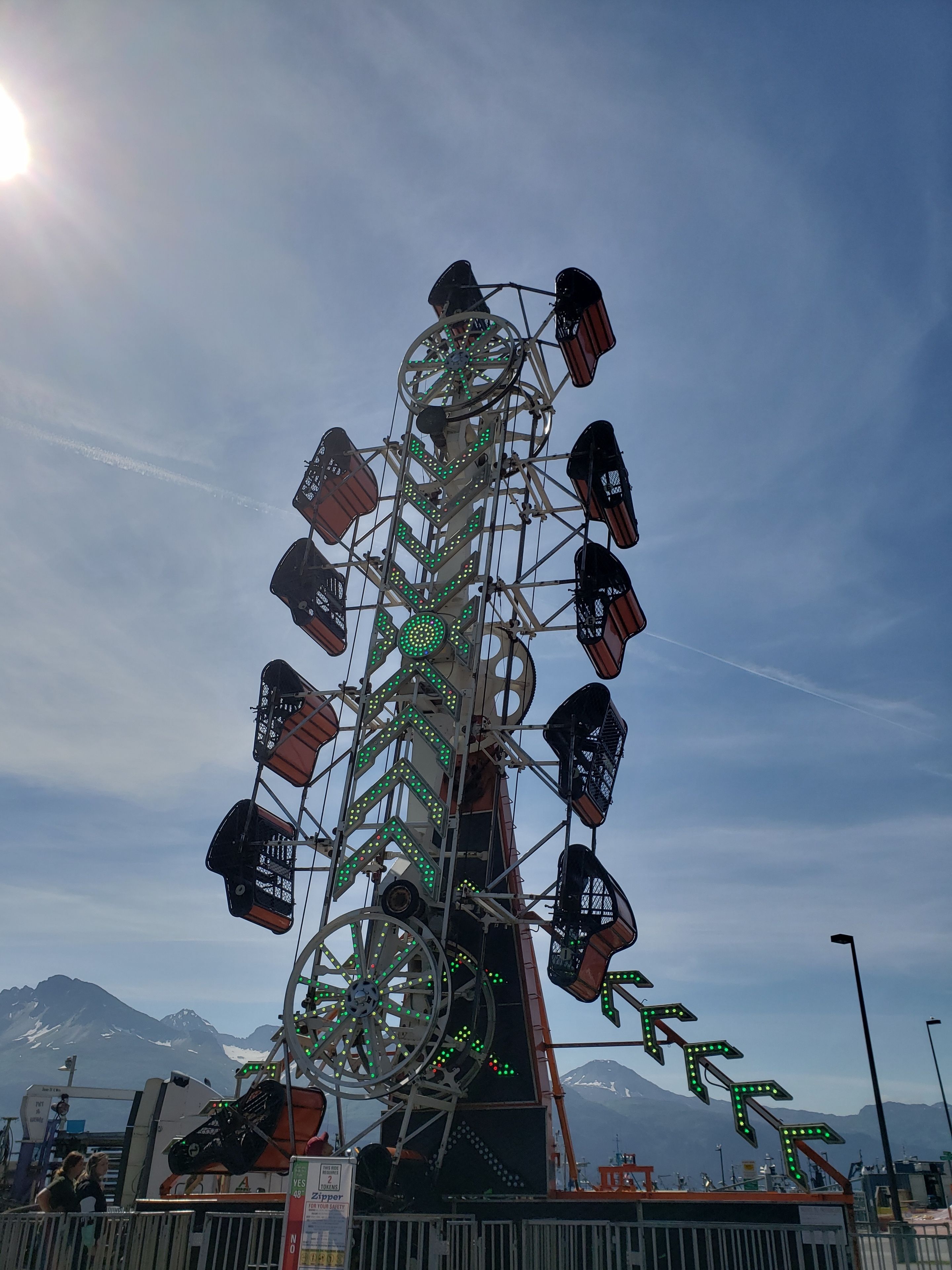 A Ferris wheel with a metal structure and green lights, set against a clear sky with mountains in the background.