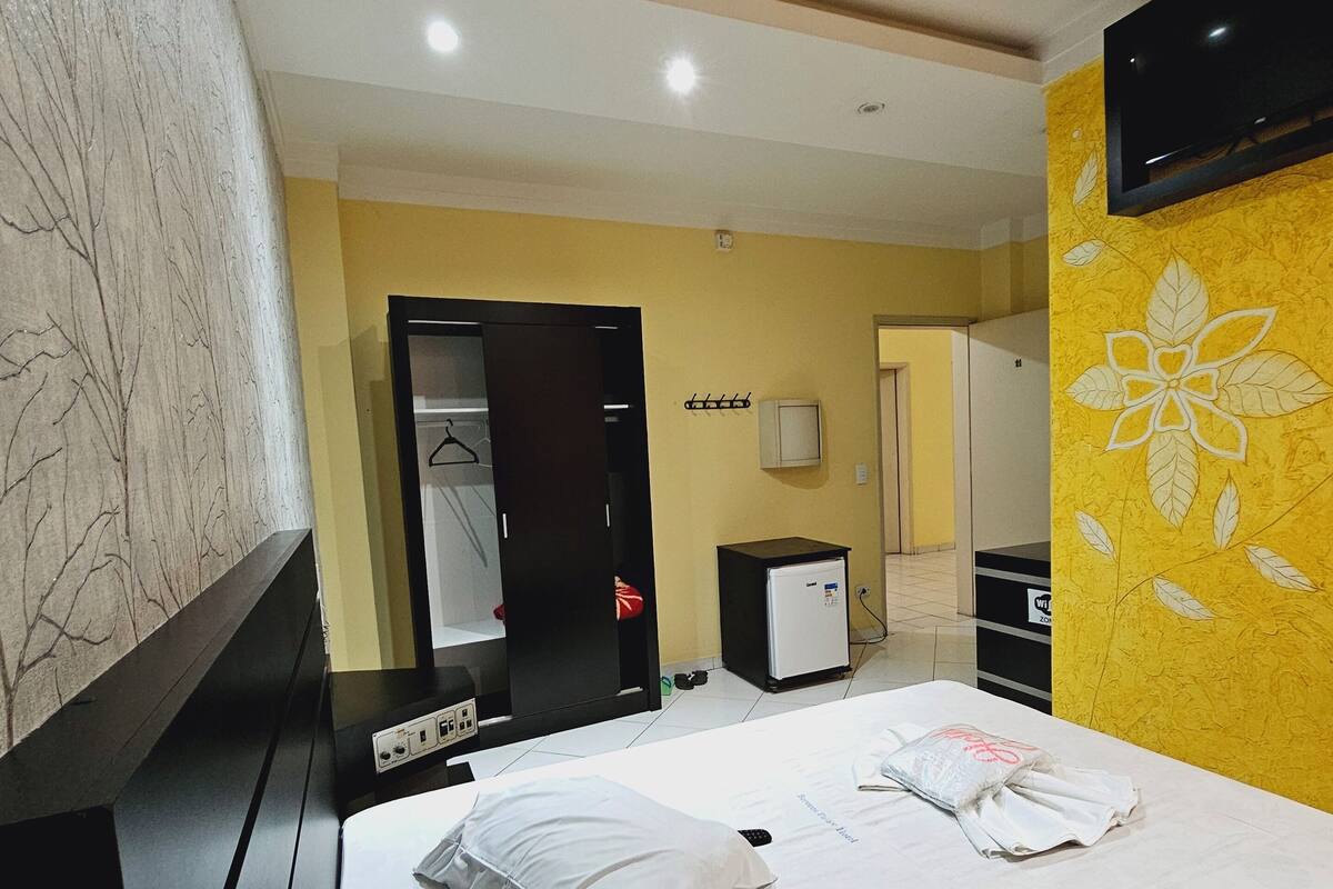 A hotel room with a bed, a TV mounted on the wall, a small refrigerator, and a coat rack.