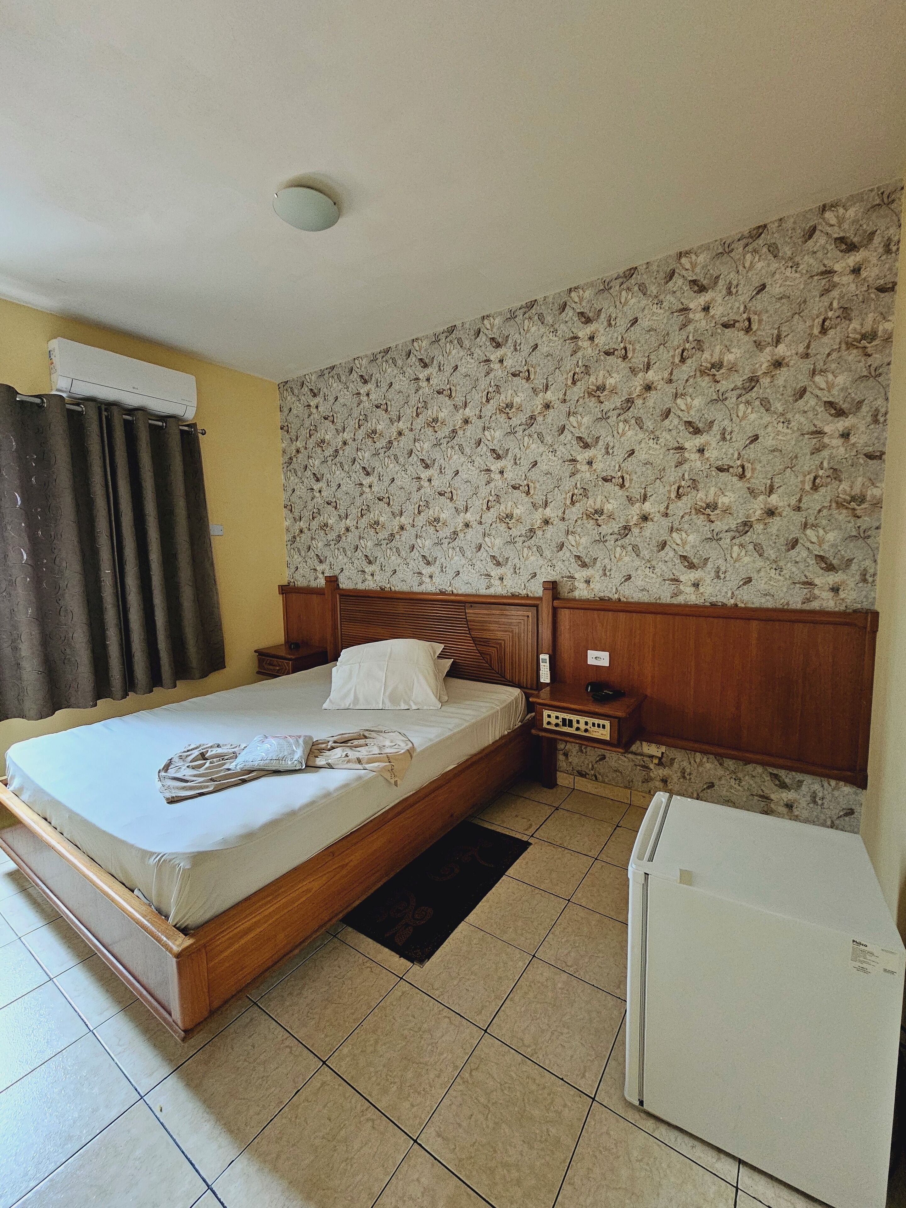 Un dormitorio con cama, mesita de noche, aire acondicionado instalado en la pared, refrigerador y un papel tapiz estampado.