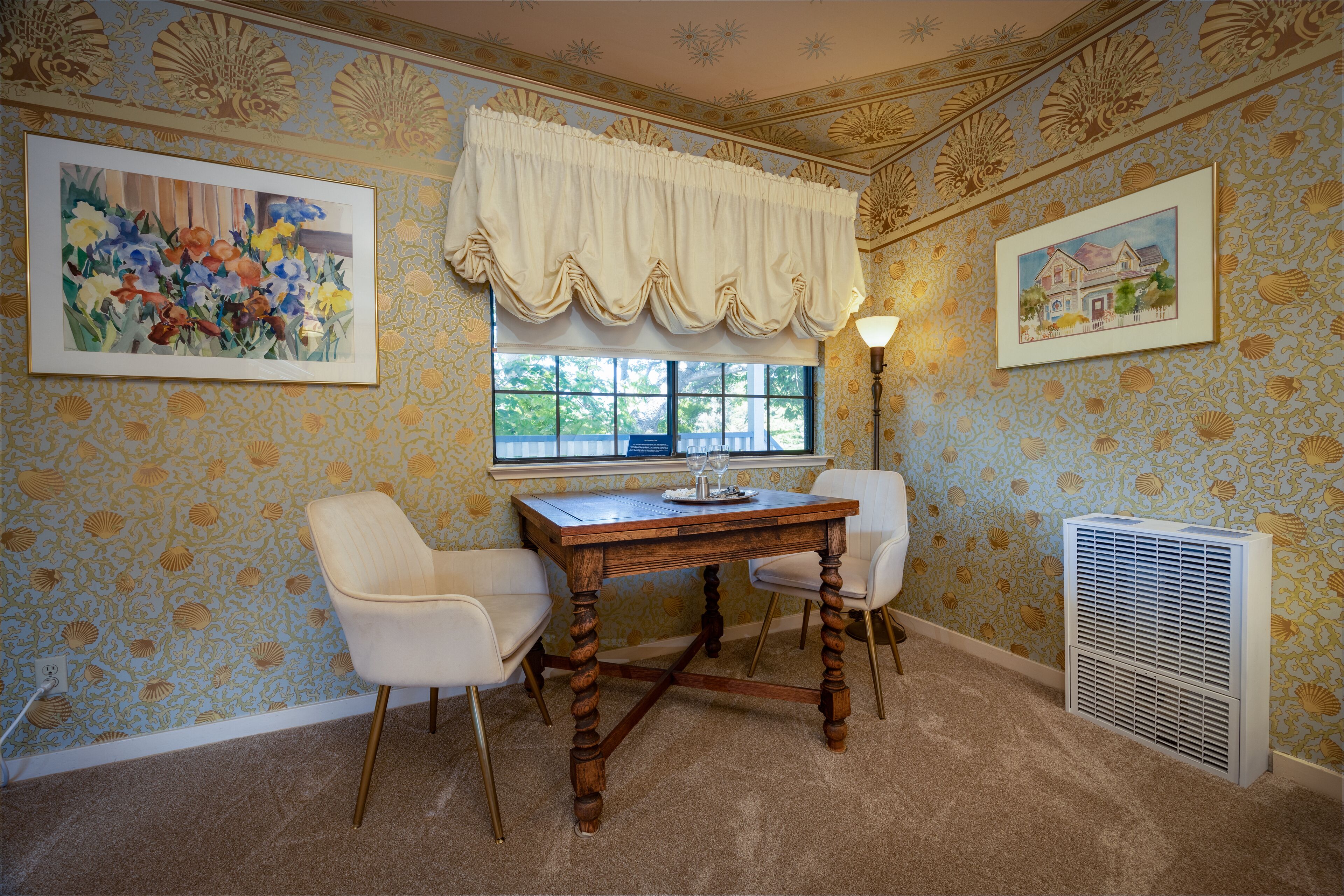 A room with a wooden table, two white chairs, a window with a valance, a wall with a floral pattern, and a radiator.