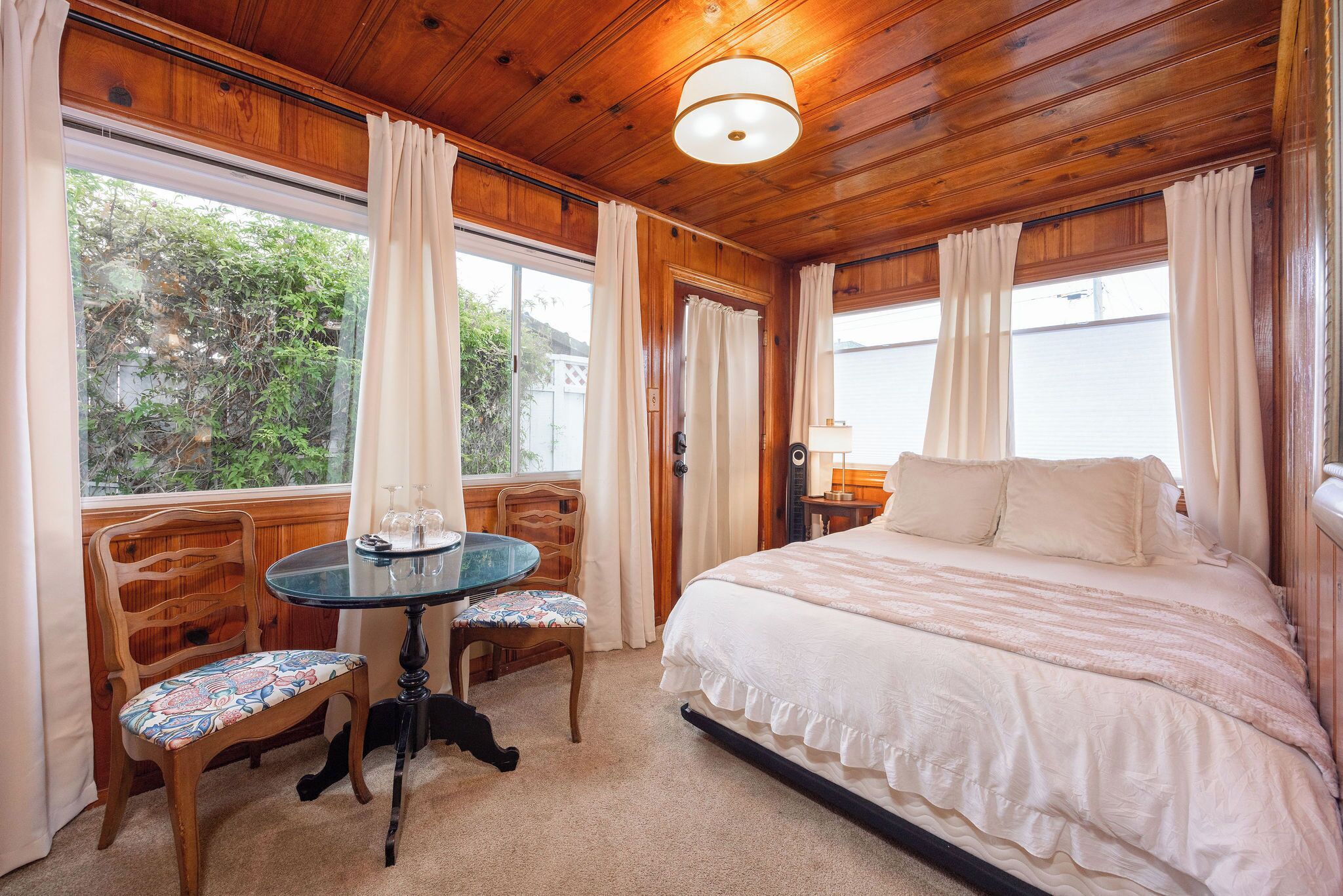 Quarto com cama, mesa redonda e cadeiras, e janela com cortinas.