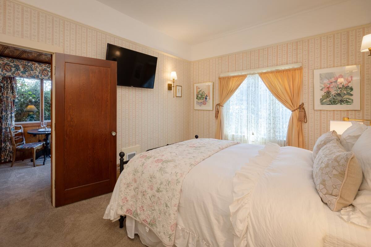 Camera da letto con un letto grande, una televisione montata a parete, un tavolino con sedie e una porta che conduce in un'altra stanza.