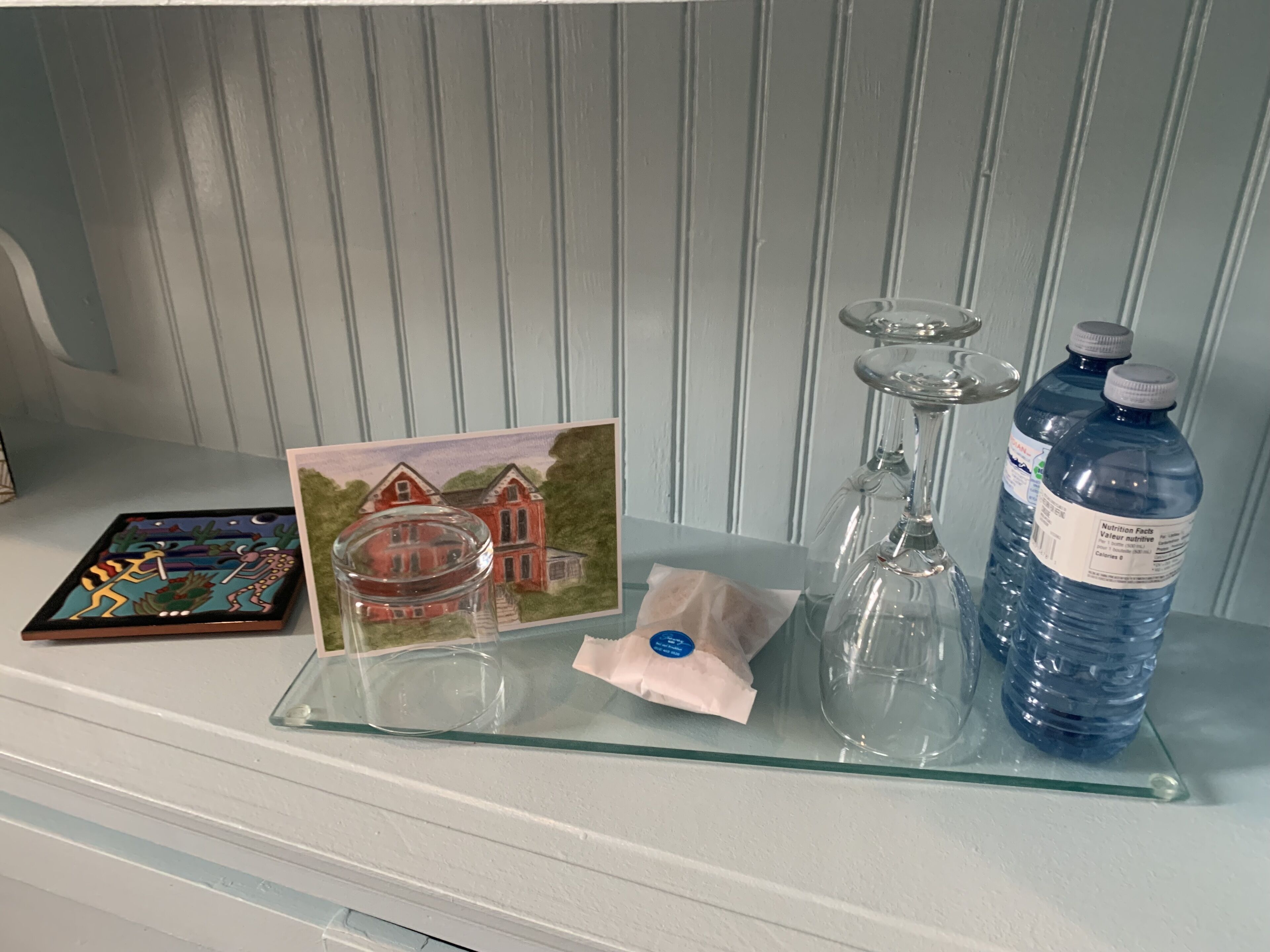 A glass tray with a glass jar, a glass candle holder, a water bottle, a card with a house illustration, and a small packet on a white shelf.