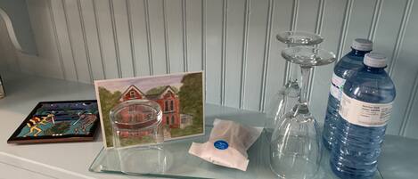 A glass tray with a glass jar, a glass candle holder, a water bottle, a card with a house illustration, and a small packet on a white shelf.