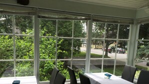 A conservatory with tables and chairs, large windows, and a view of the street and greenery outside.