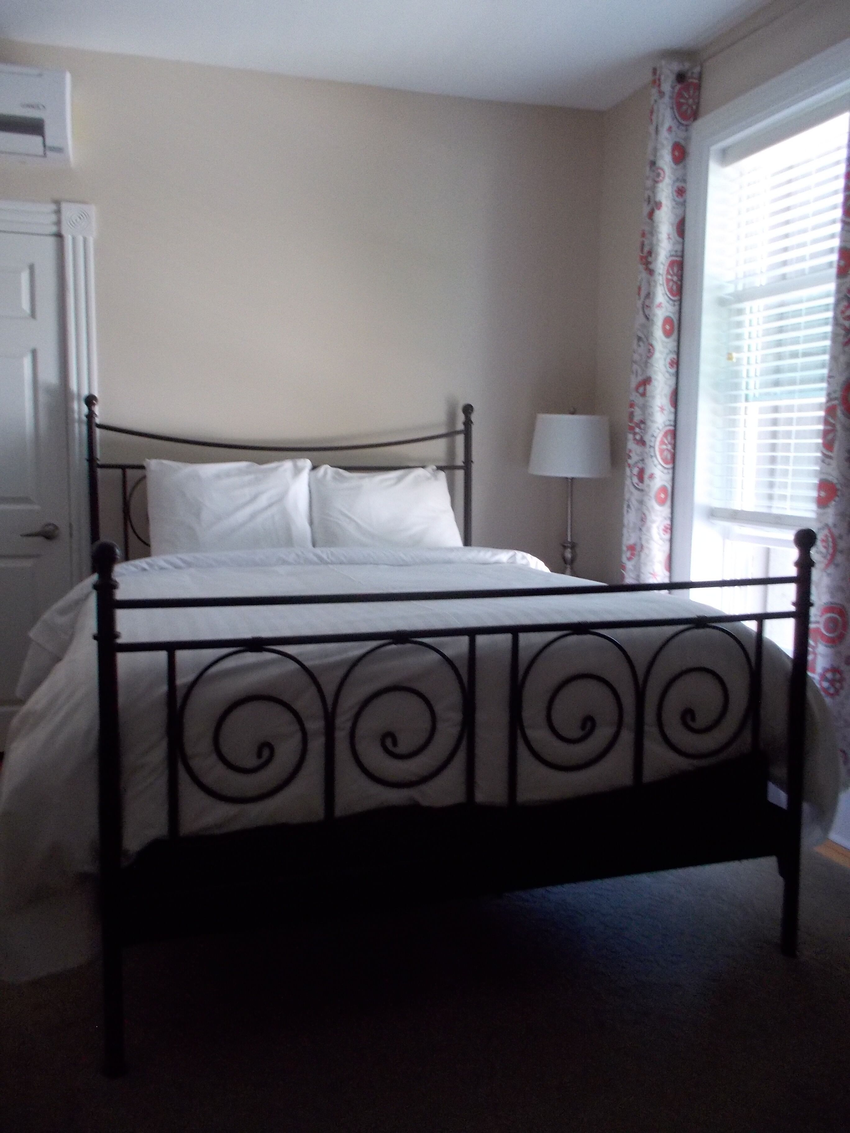 Una cama con estructura metálica y ropa de cama blanca en una habitación con ventana y puerta.