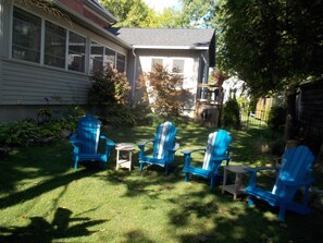 Un jardin avec une pelouse, quatre chaises Adirondack bleues, une petite table et une maison dotée d’une fenêtre et d’une porte.
