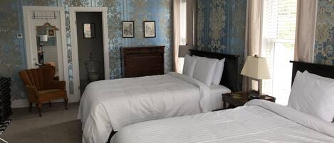 A hotel room with two beds, a wooden chair, a bedside table with a lamp, and a wardrobe.