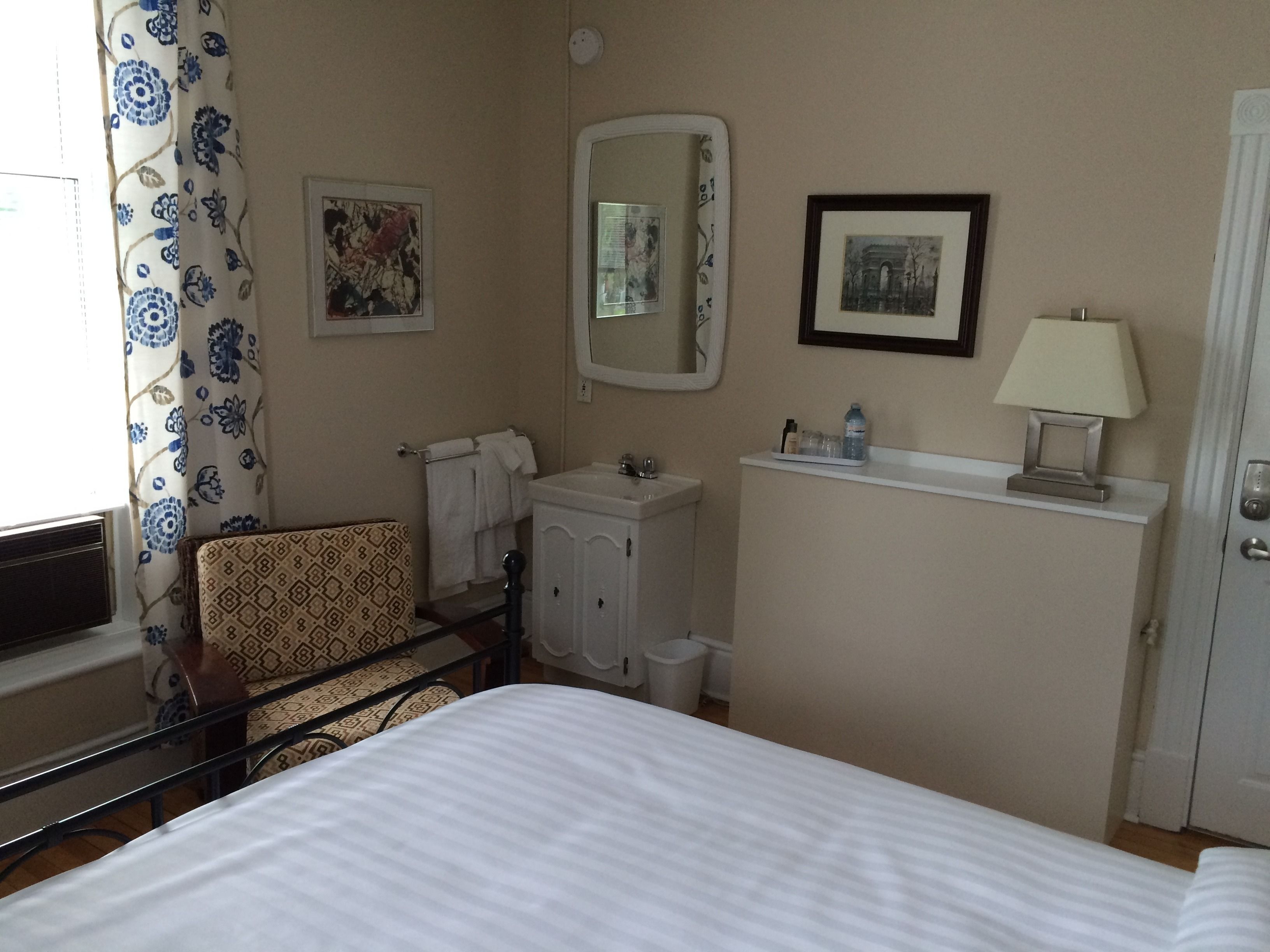 Une chambre d’hôtel avec un lit, une chaise, un lavabo, un miroir et un tableau encadré au mur.