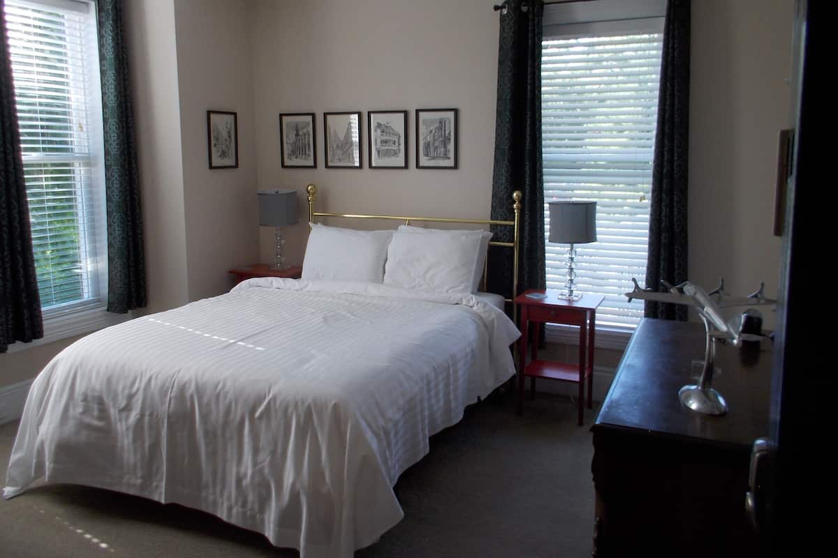 A neatly made bed with white linens, a bedside table with a lamp, and a chair. There are four framed pictures on the wall above the bed.